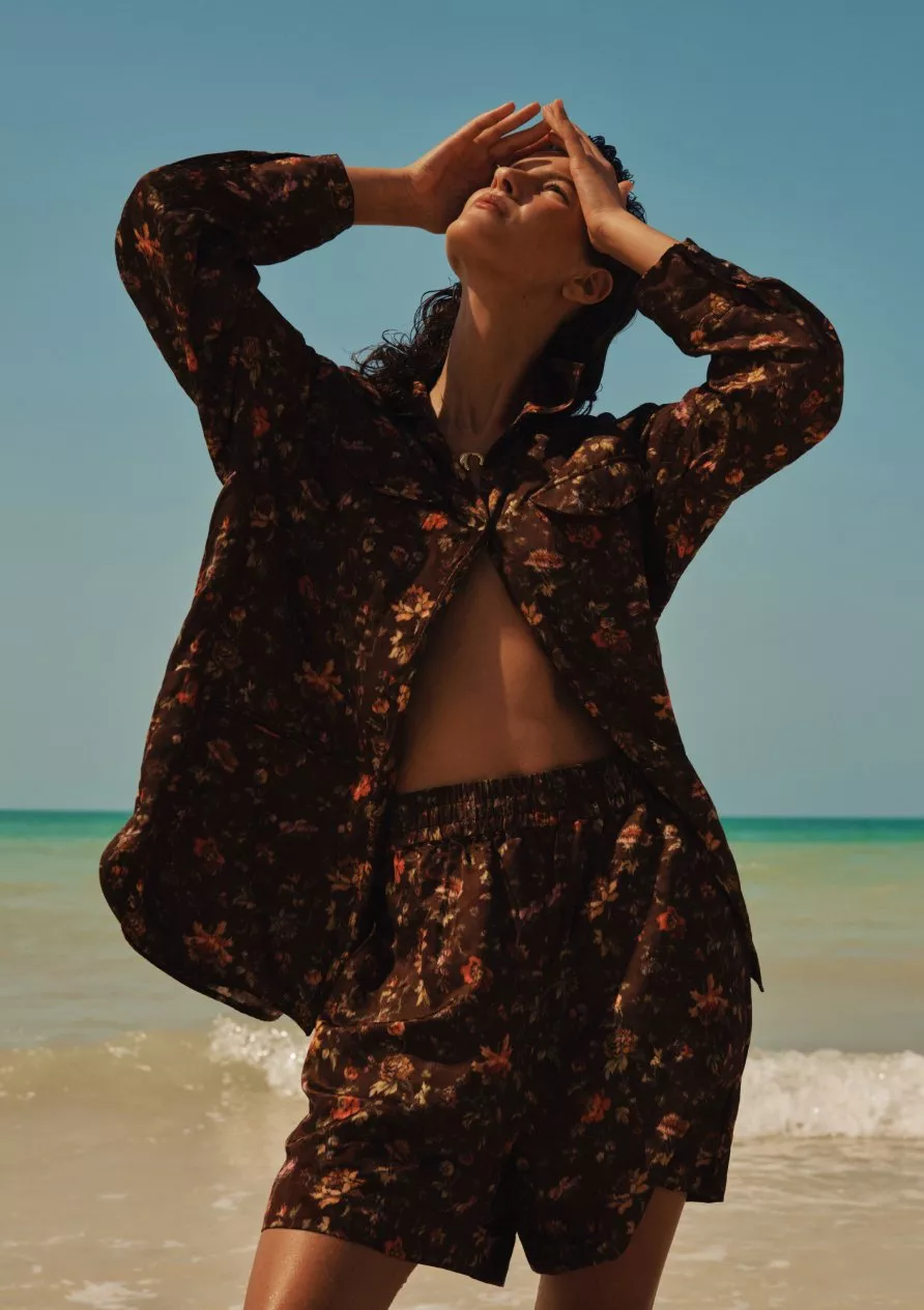 Fashion model on sunny beach wearing floral shirt and matching shorts, hands raised, ocean and blue sky backdrop