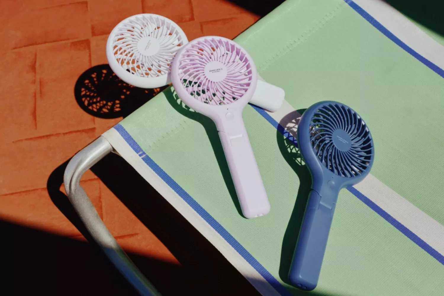 Three portable handheld mini fans on a green striped sun lounger, summer cooling gadgets in bright sunlight
