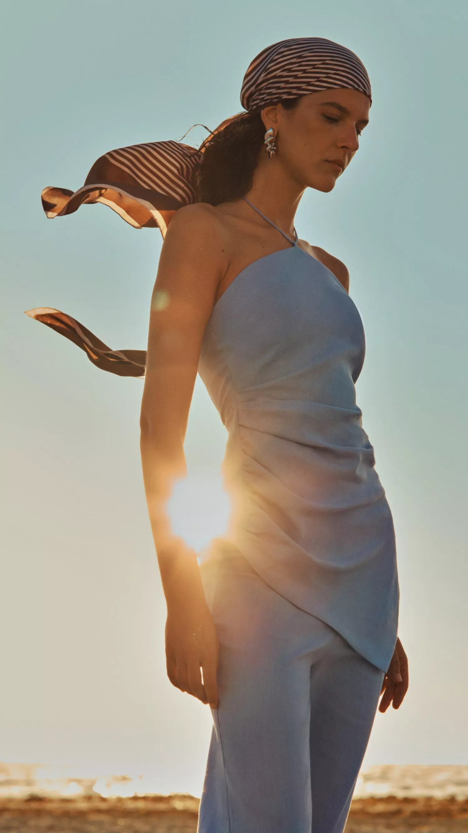 Fashion model in pale blue slip dress and striped headscarf, backlit by sunset on a beach shoreline