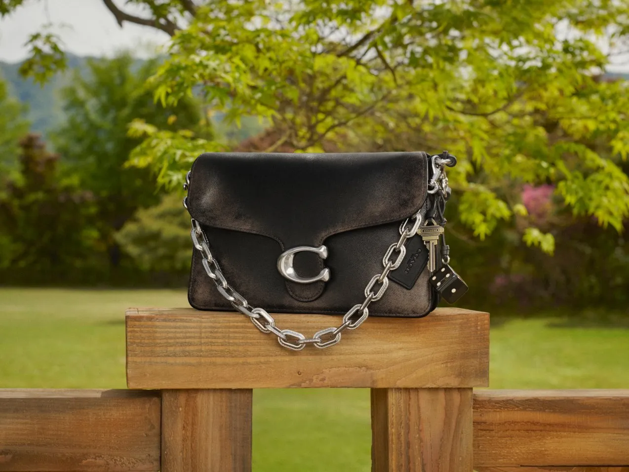 Black leather shoulder bag with silver chain strap and C clasp, displayed outdoors on a wooden rail in a garden.