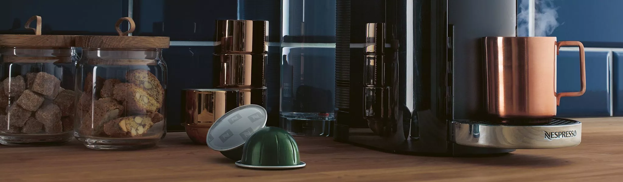 Nespresso coffee machine brewing espresso beside copper mug, coffee pods and biscuit jars on kitchen worktop