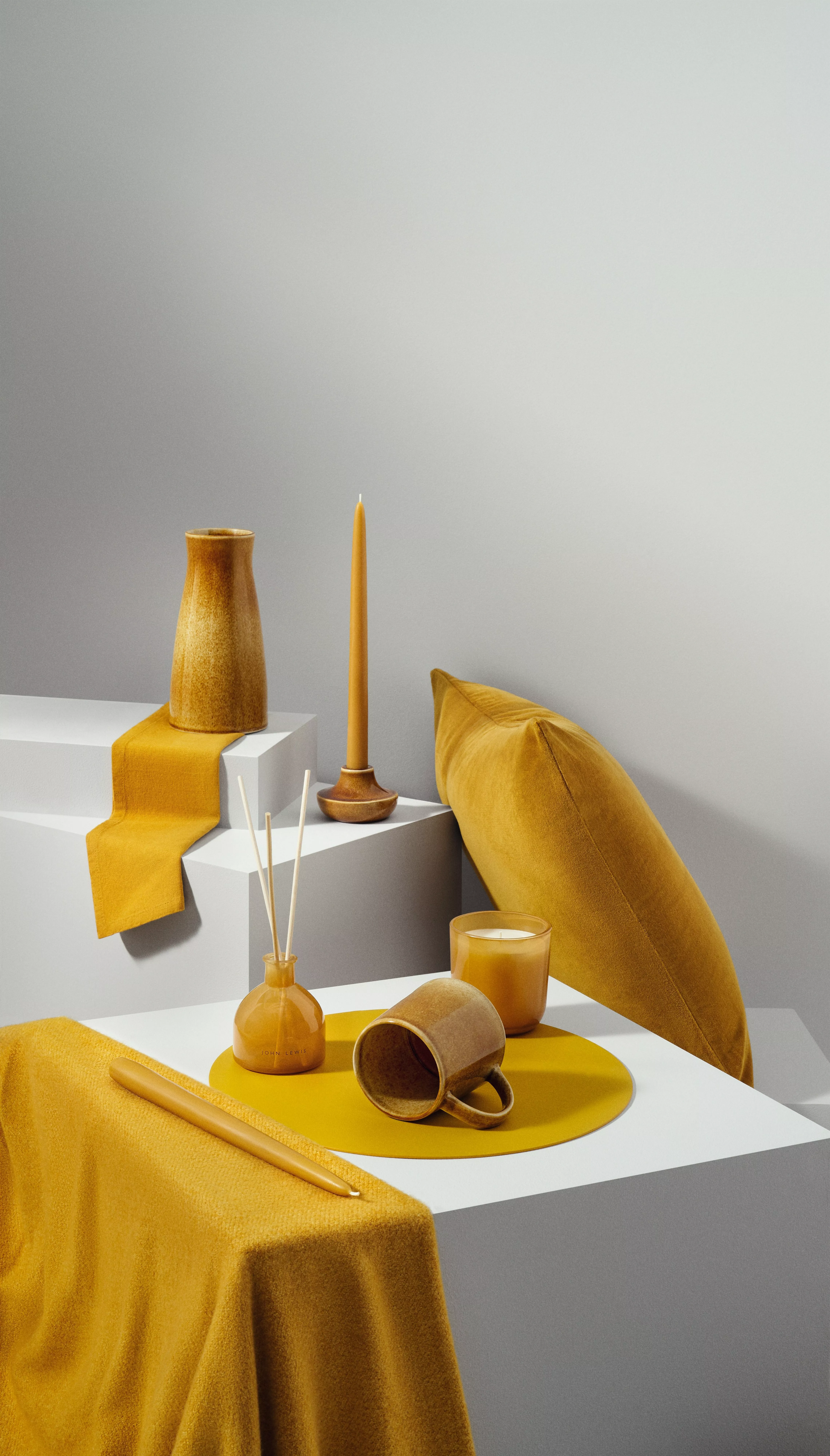 Mustard yellow home décor still life with ceramic vase, candle, cushion, mug and reed diffuser on white blocks