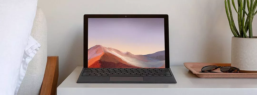 Laptop on bedside table with mountain wallpaper, beside sunglasses on tray and potted plant in modern bedroom