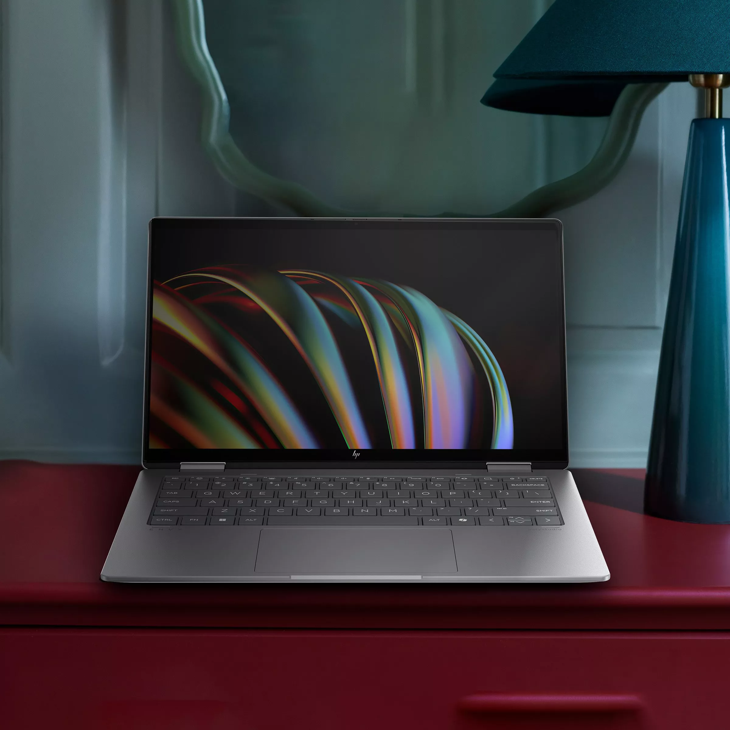 HP laptop on a red desk with colourful abstract wallpaper, beside a blue lamp in a modern home office setting