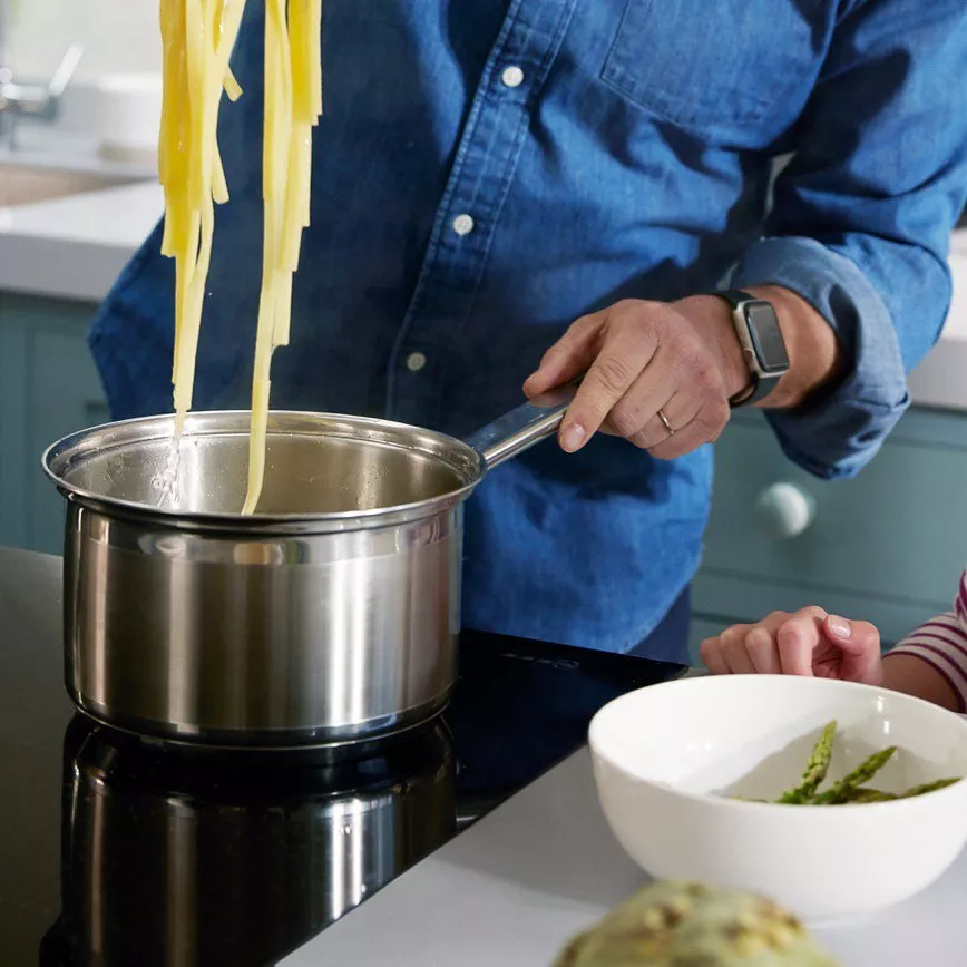 Person in denim shirt cooking pasta in a stainless steel pot on a stove, with asparagus nearby.