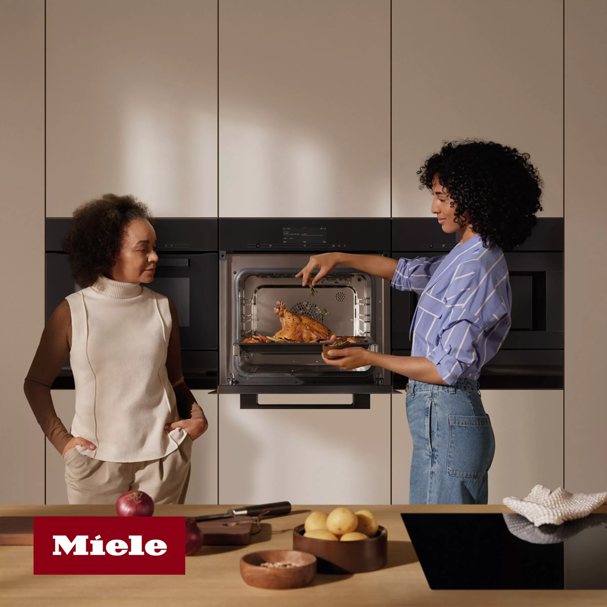 Person putting a turkey in the oven, next to another person standing by the kitchen counter