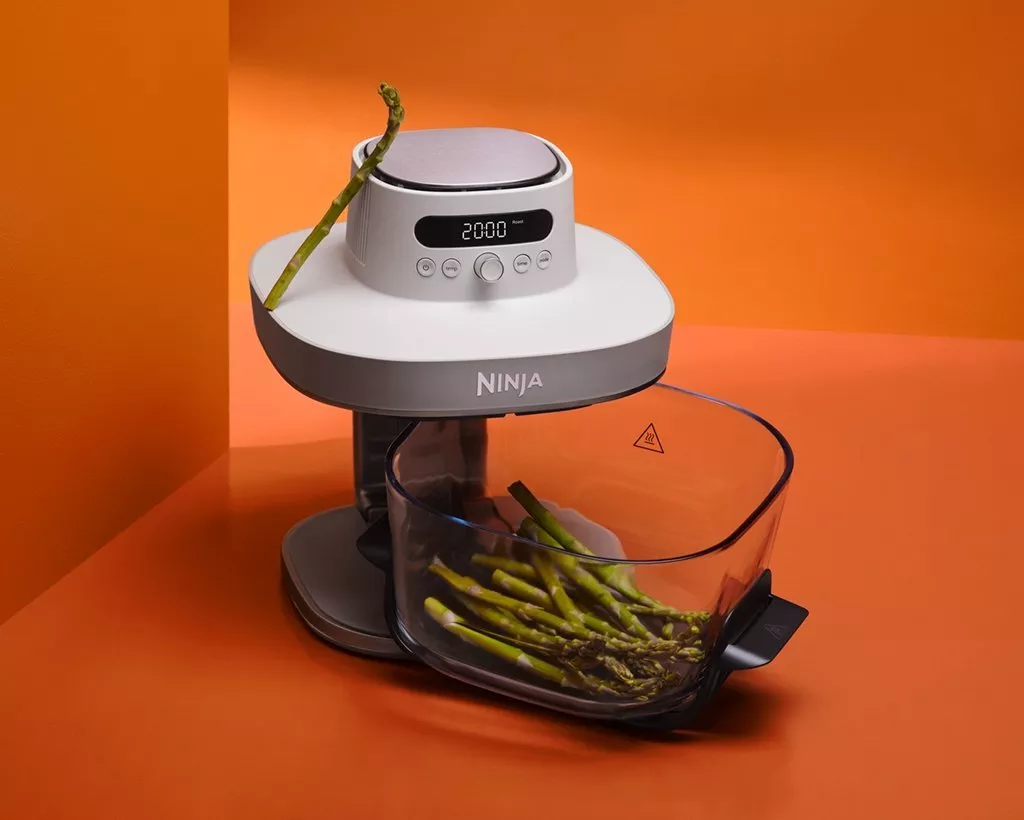Ninja food processor with digital timer, clear bowl of fresh asparagus spears, on bright orange background.