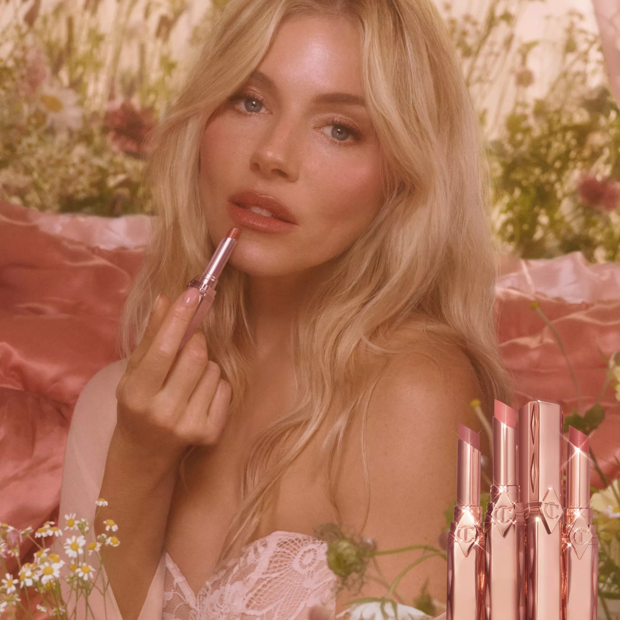 A woman with blonde hair applies lipstick in a floral setting. Four lipsticks are displayed in the foreground.