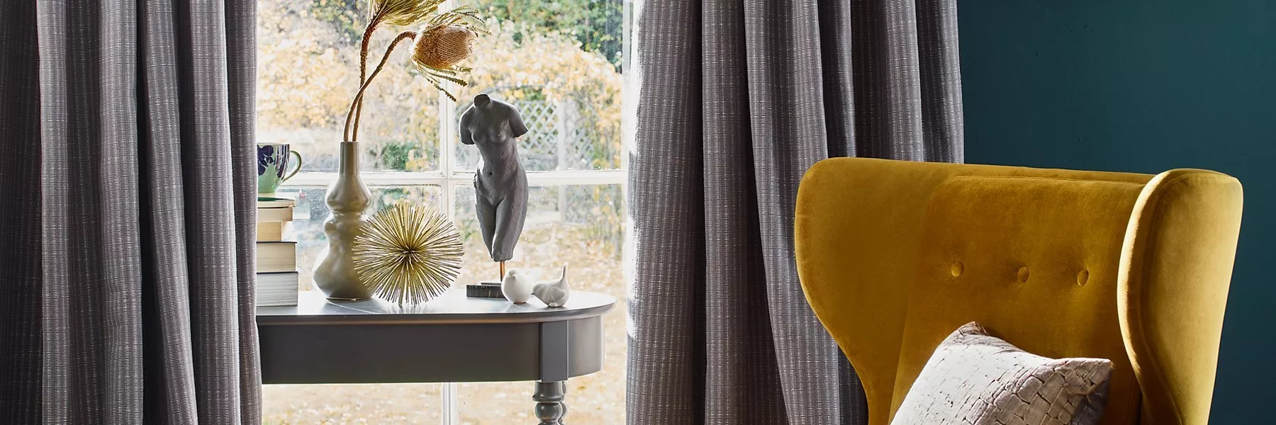 Grey textured curtains framing a window, with a mustard velvet armchair and modern decorative table display.