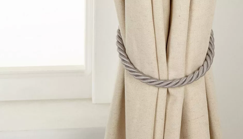 Close-up of beige curtain with grey twisted rope tieback beside a white window frame, home décor detail