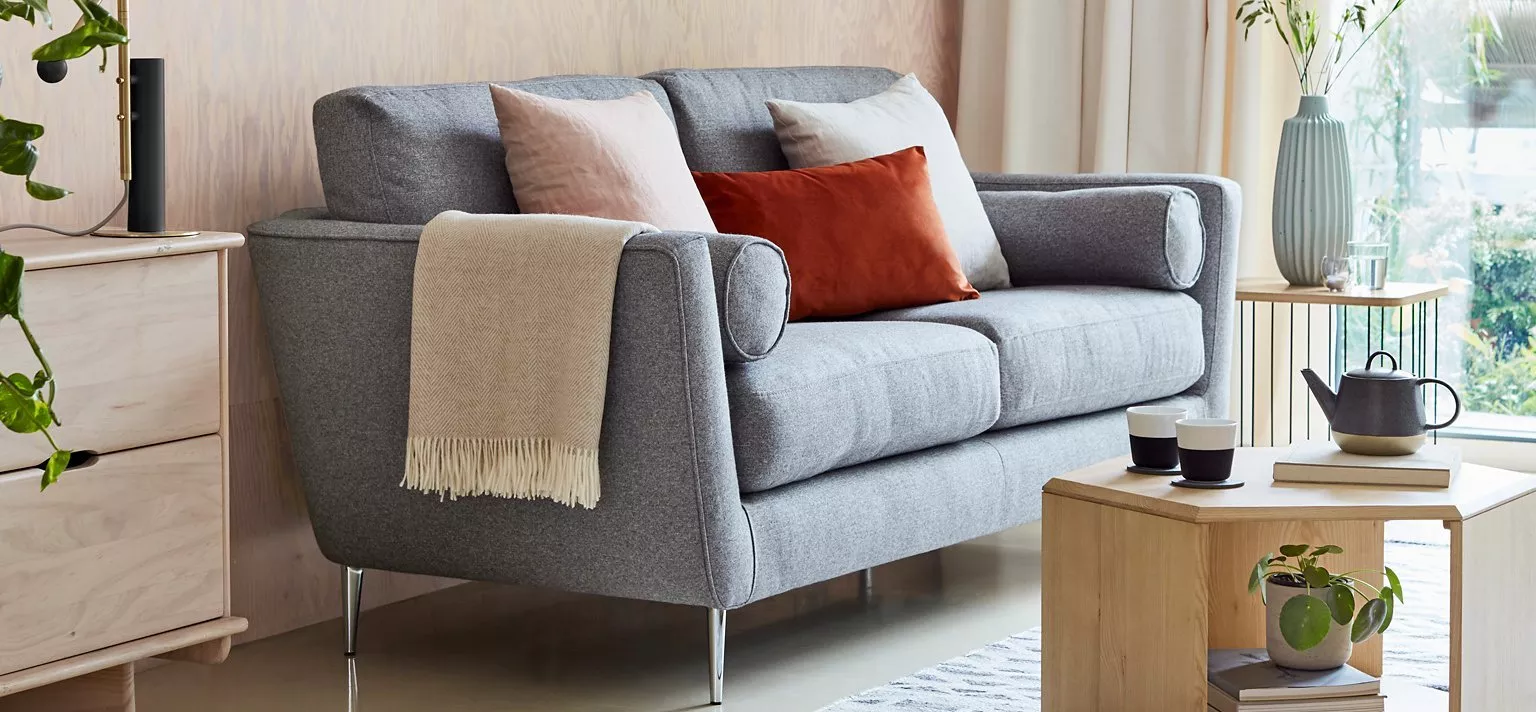 Modern grey fabric sofa with cushions and throw in cosy living room, wooden coffee table and tea set near window