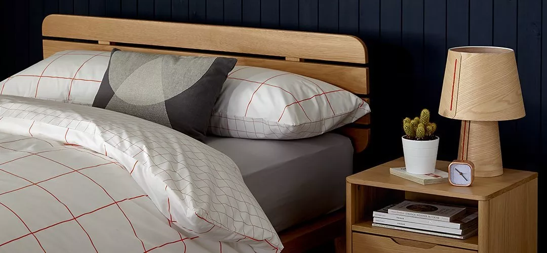 Modern wooden bed with white patterned bedding and oak bedside table with lamp, cactus plant and clock