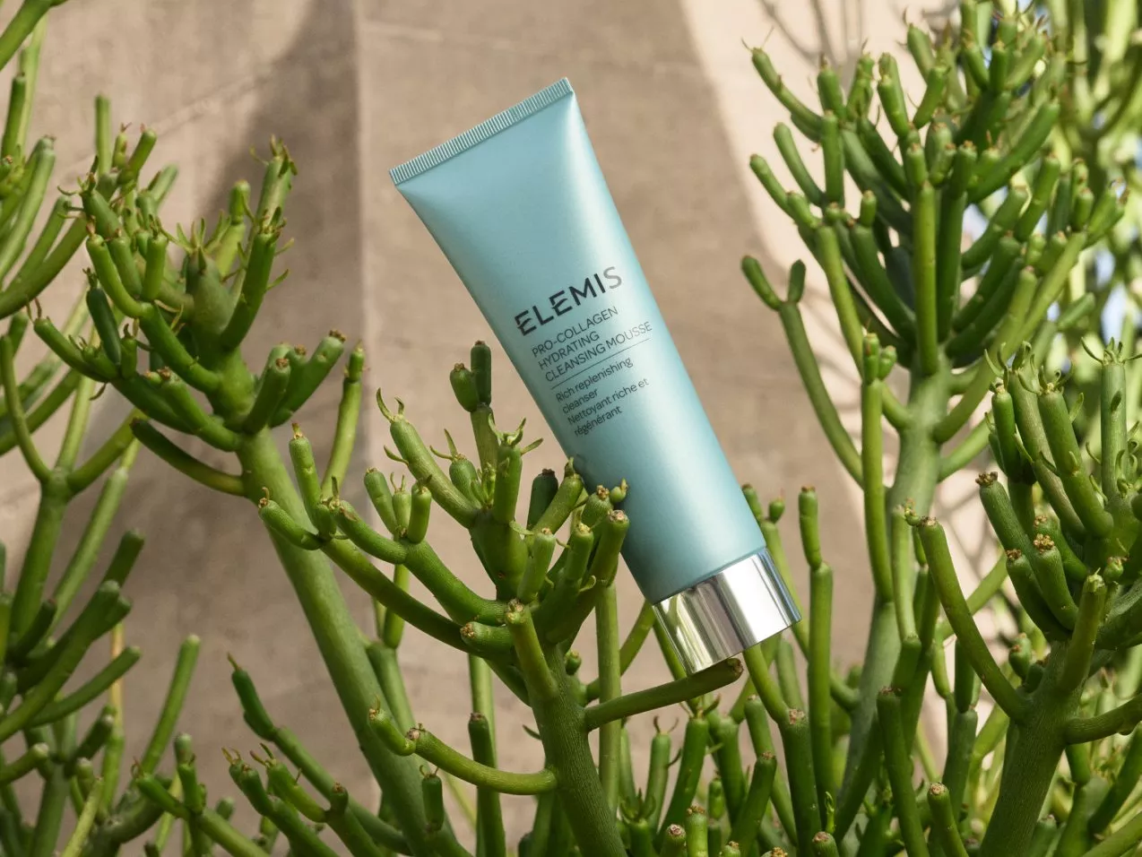 A blue tube of Elemis Pro-Collagen Cleansing Mousse is nestled among vibrant green plants outdoors.