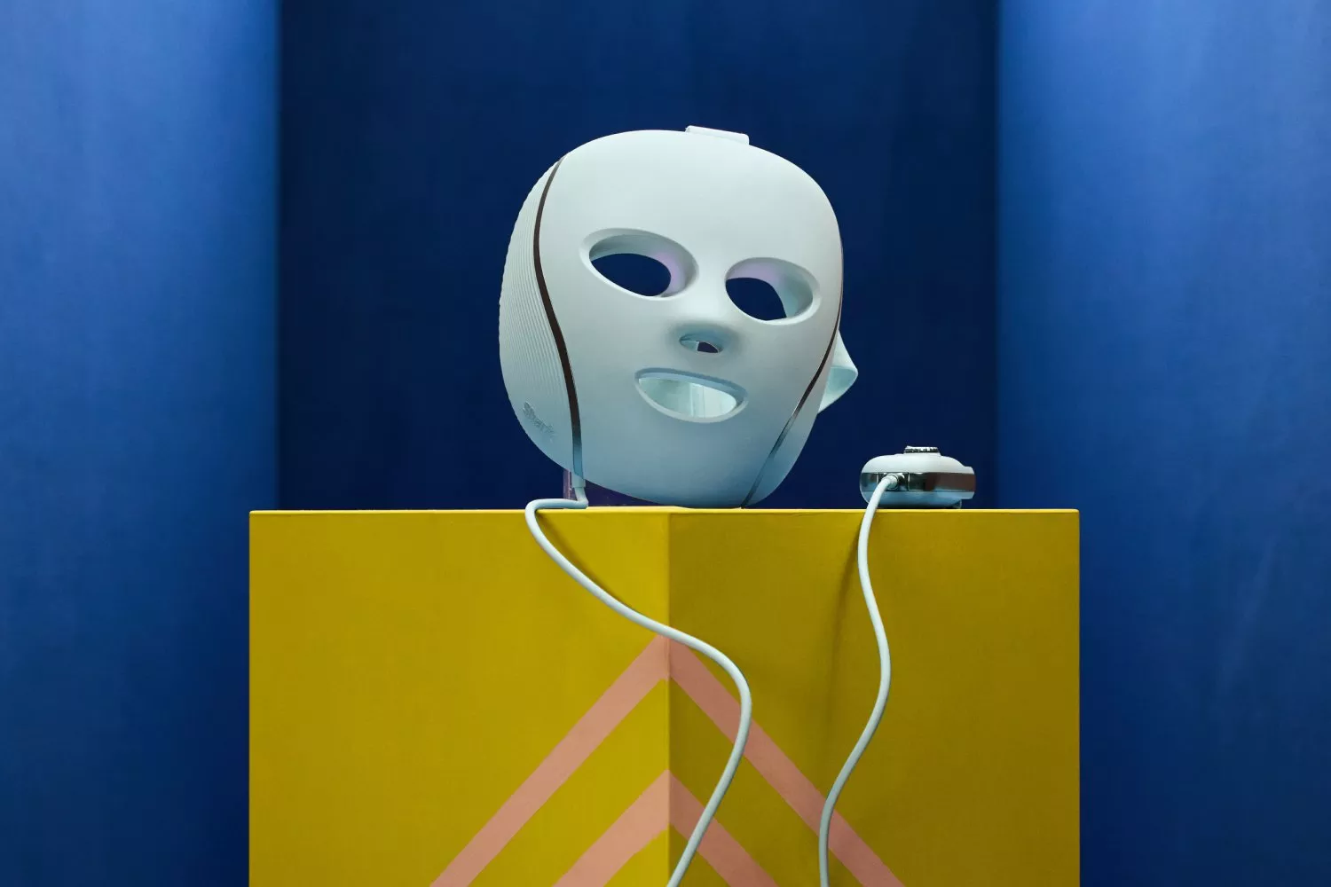 A white robotic device with cables displayed on a bright yellow stand with geometric patterns and a blue backdrop.