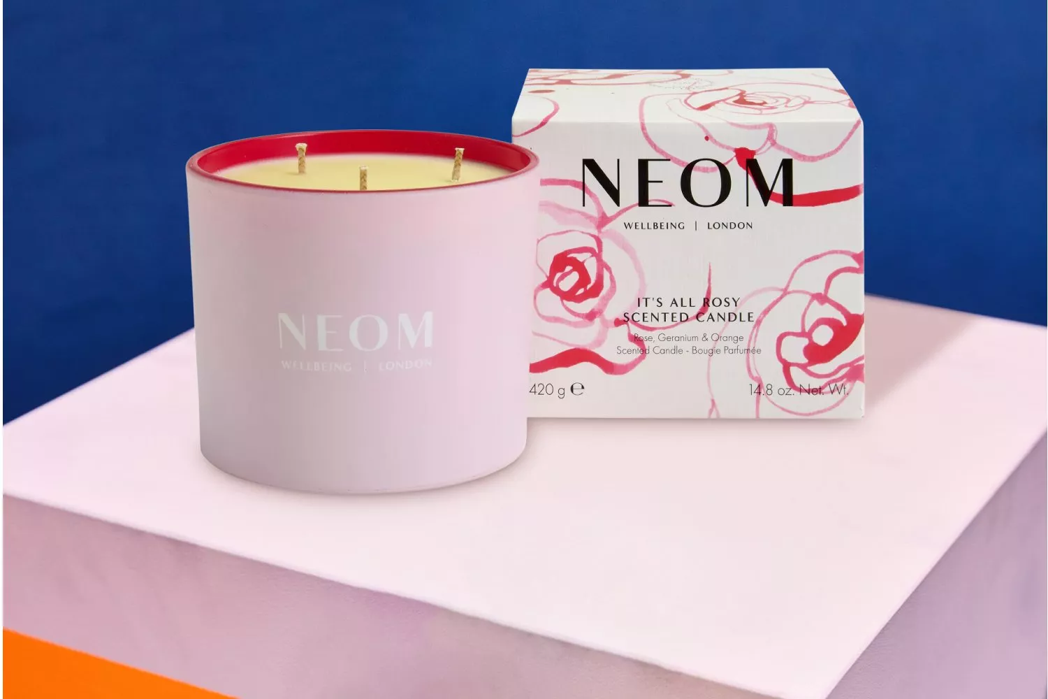 NEOM scented candle and box on colourful surface. Candle is pink, box has floral design, text "It's All Rosy".
