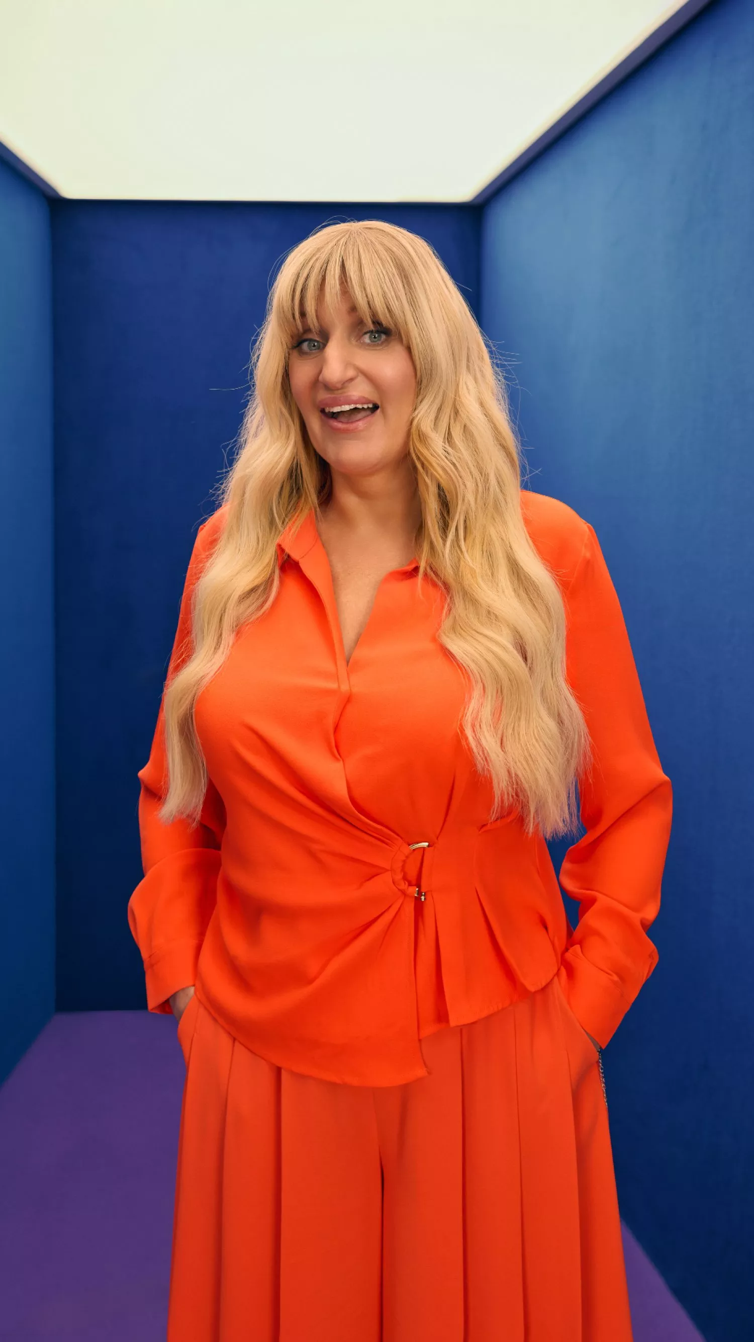 A person with long blonde hair wears a stylish bright orange outfit against a blue geometric backdrop.