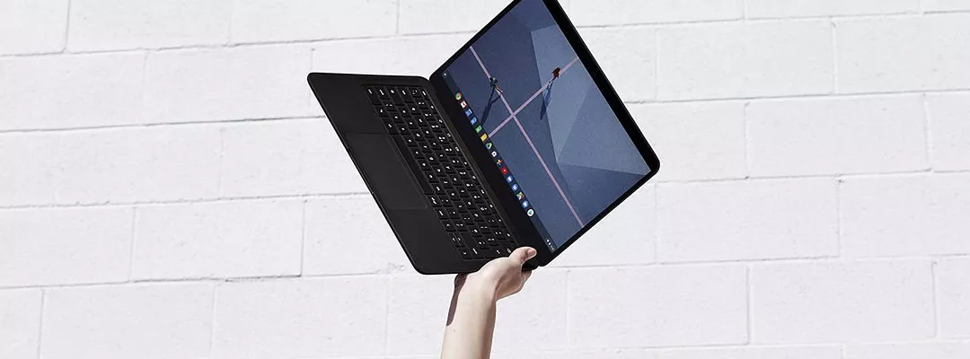 Hand holding a sleek black laptop with touchscreen, tilted open against a white brick wall background