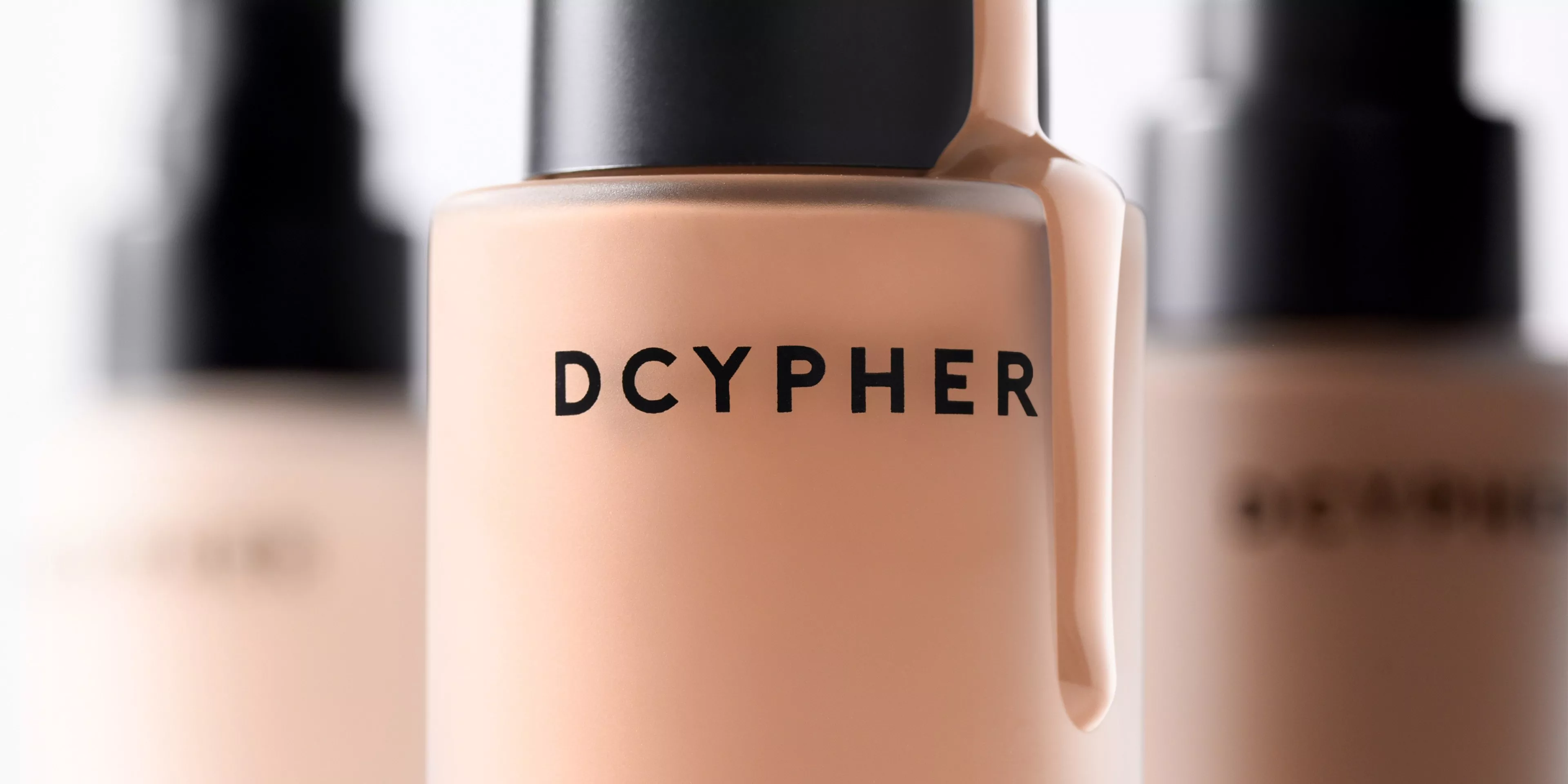 Close-up of DCYPHER liquid foundation bottle with black cap, beige makeup shade, blurred bottles behind