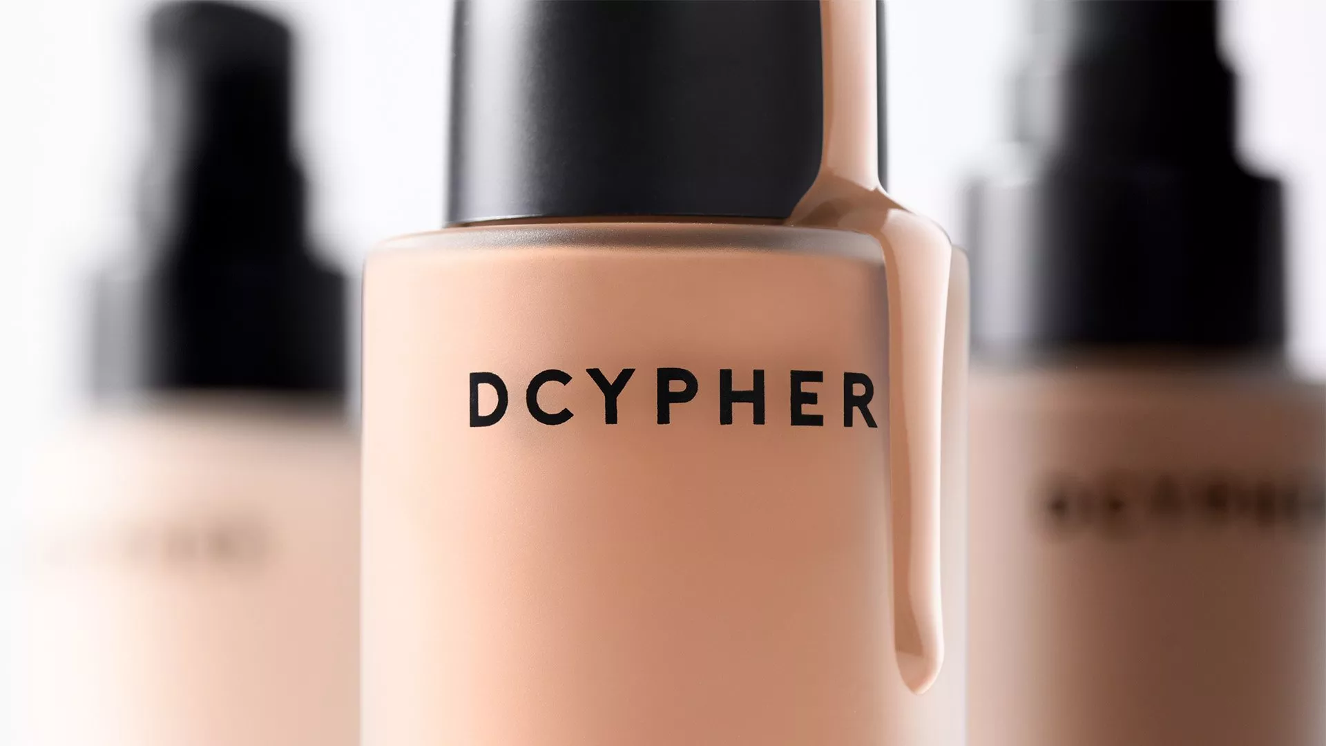 Close-up of DCYPHER liquid foundation bottle with black cap, beige makeup shade, blurred bottles behind
