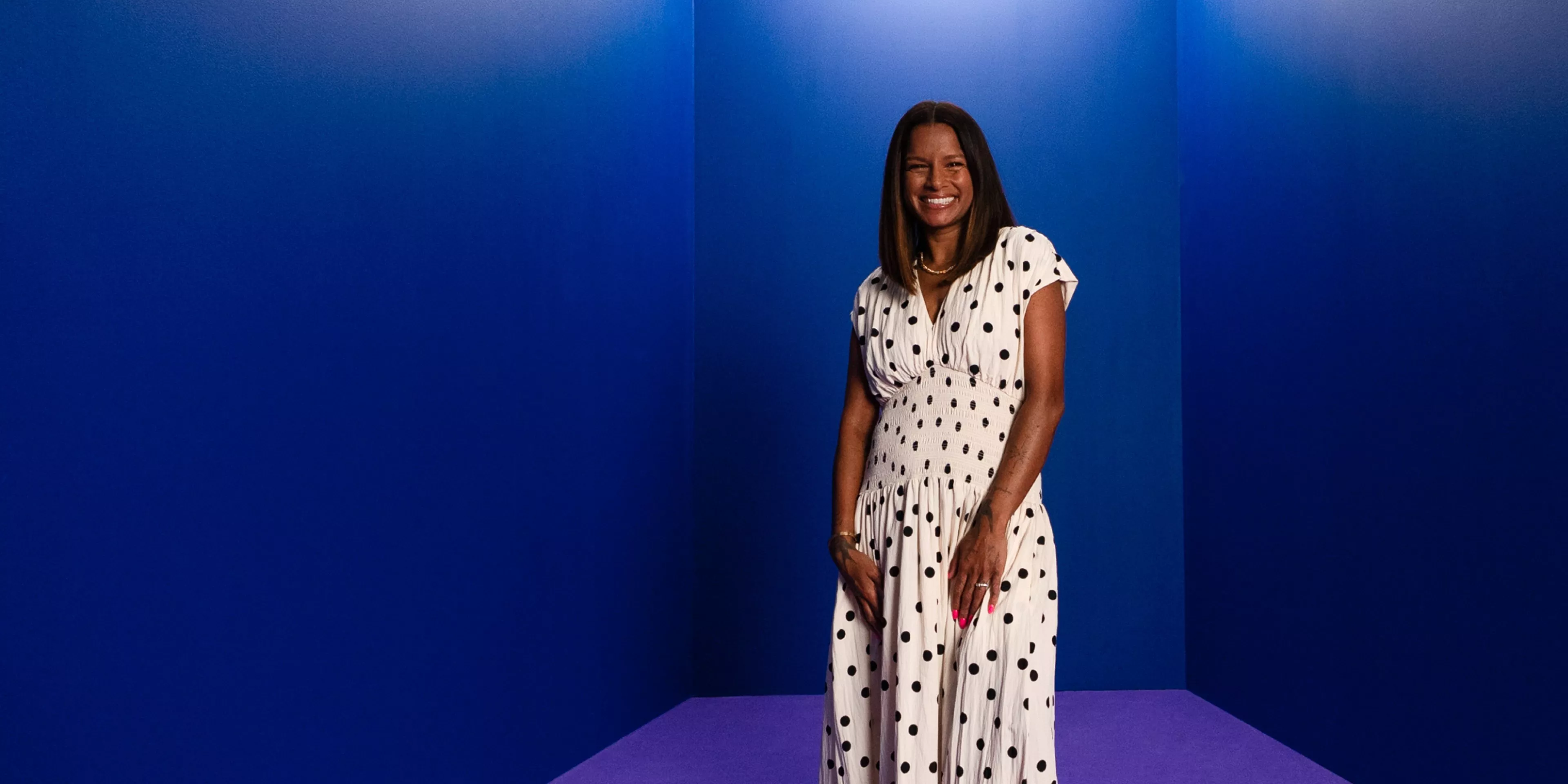 Smiling woman in white polka dot dress standing in a blue studio corner, full-length fashion portrait.