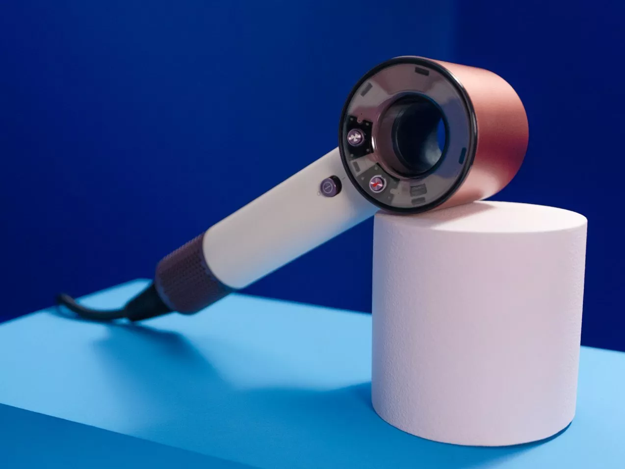 Modern hair dryer on white plinth against blue backdrop, featuring rose gold barrel and sleek ergonomic handle