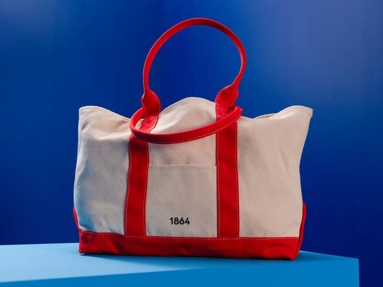 Beige canvas tote bag with red handles and trim, “1864” logo, displayed on blue pedestal background