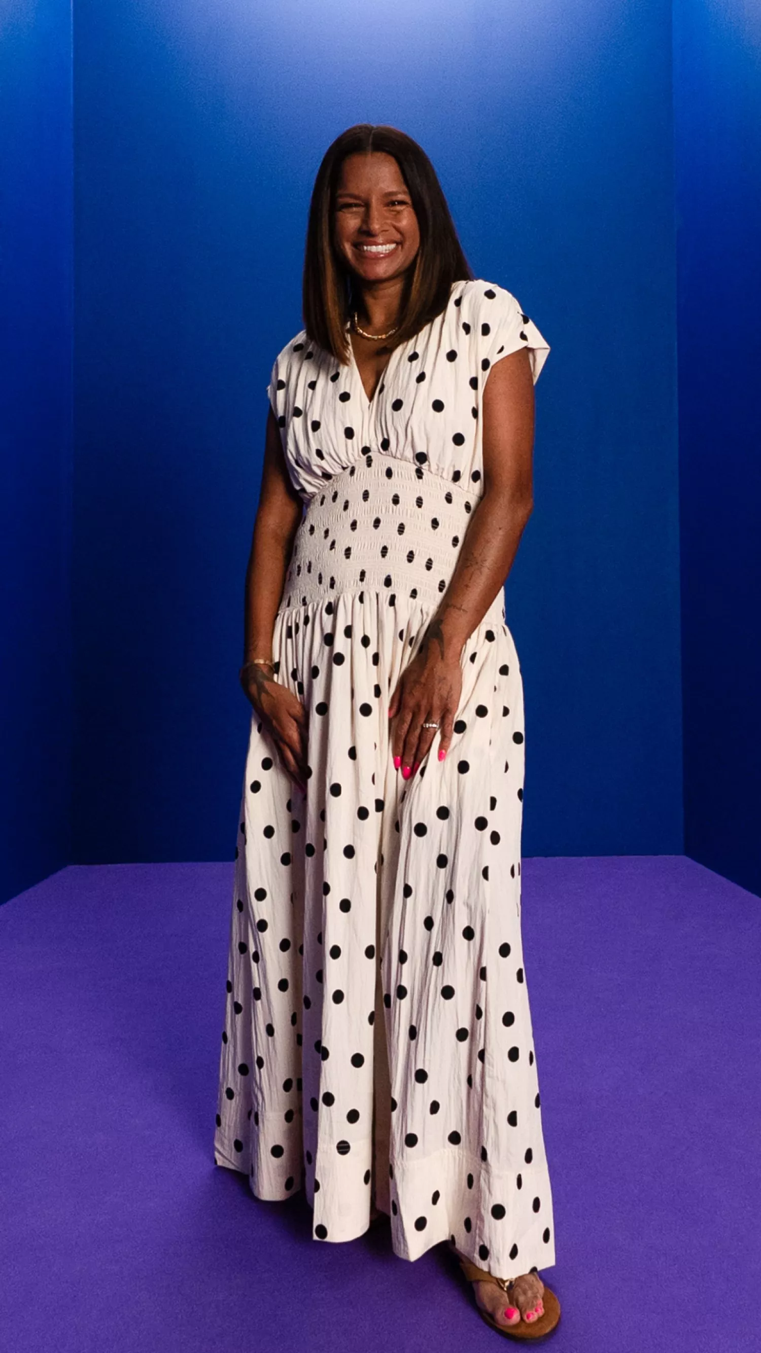 Smiling woman in white polka dot maxi dress posing in studio with blue walls and purple floor backdrop