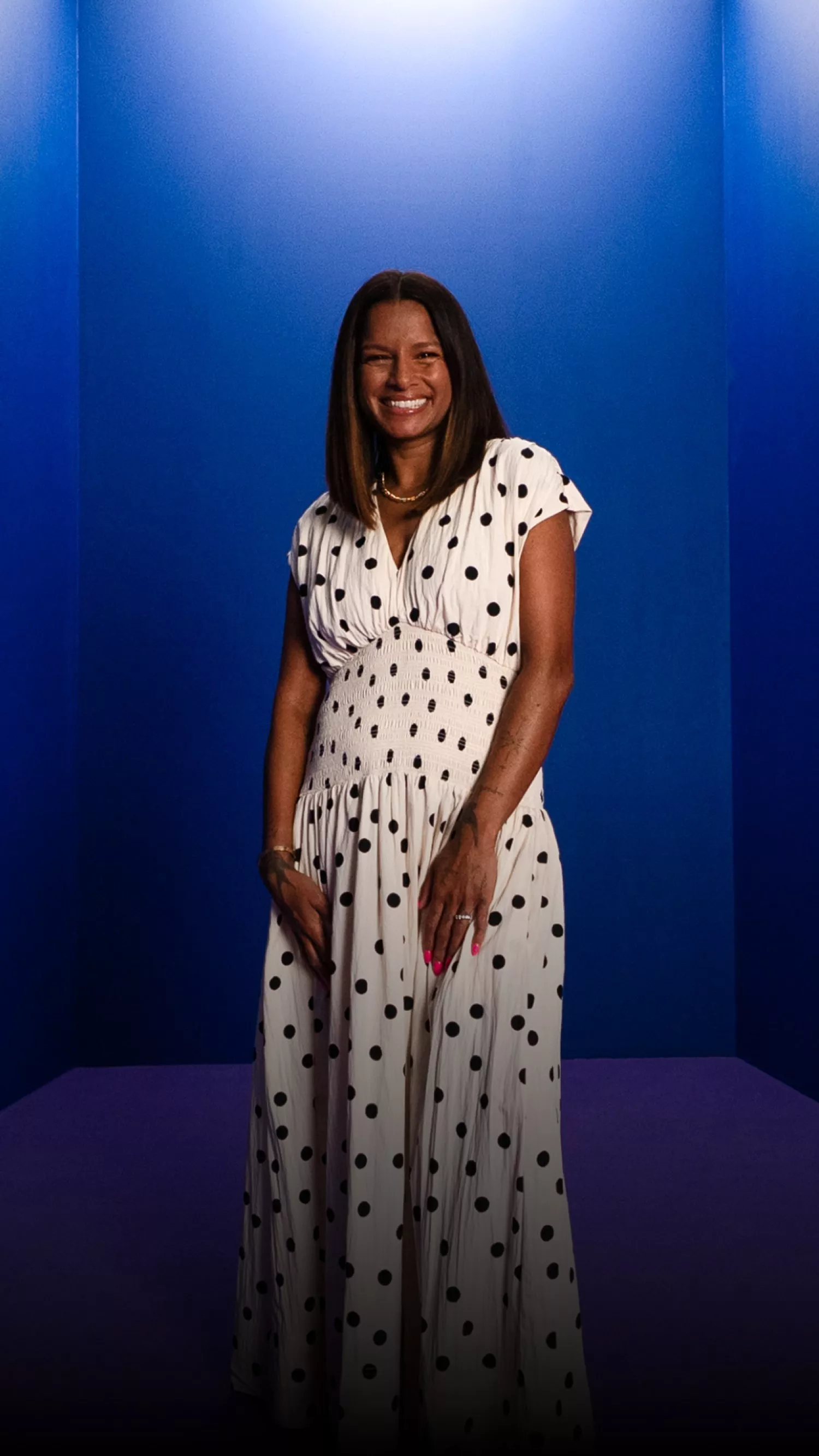 Smiling woman in white polka dot maxi dress posing in studio with blue walls and purple floor backdrop