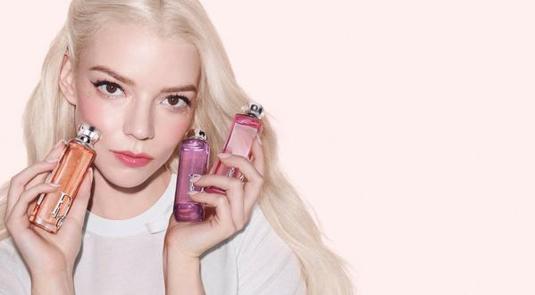 Women with long blonde hair holds two perfume bottles, one orange and one pink, against a soft pink background.