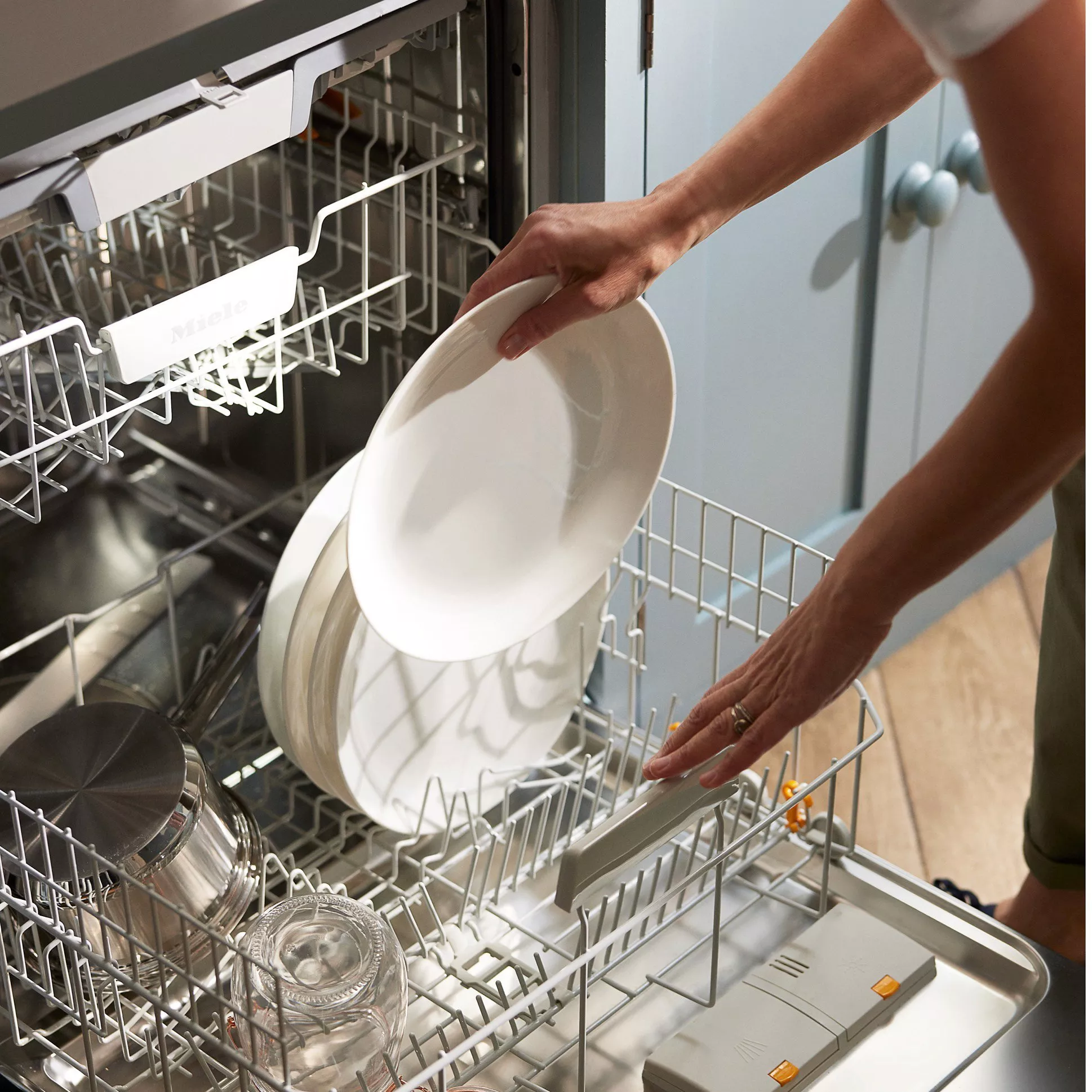 Person loading white plates into a dishwasher with stainless steel pots and clear glasses inside.