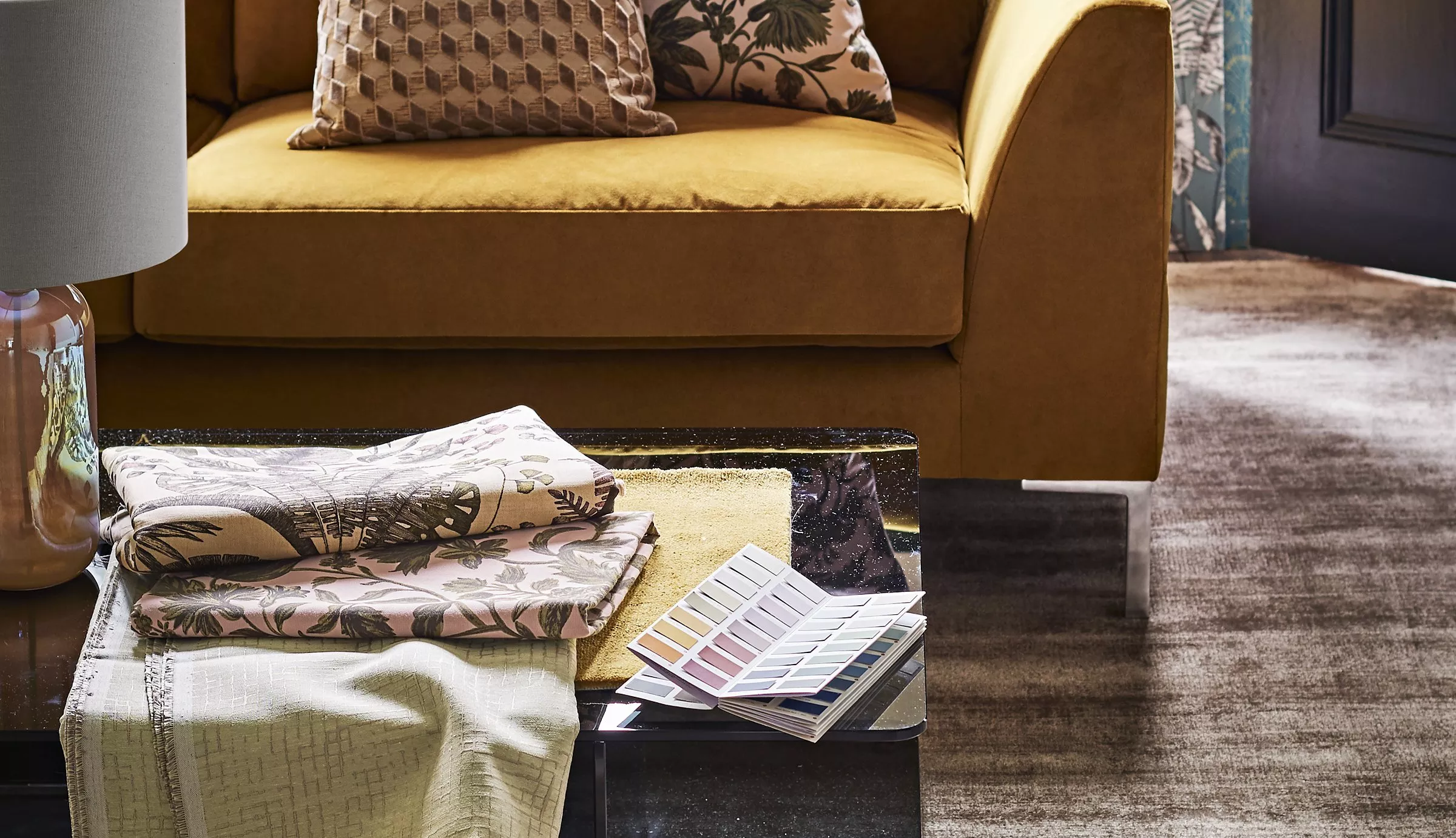 Mustard velvet sofa with patterned cushions, fabric samples and colour swatches on glass coffee table in lounge