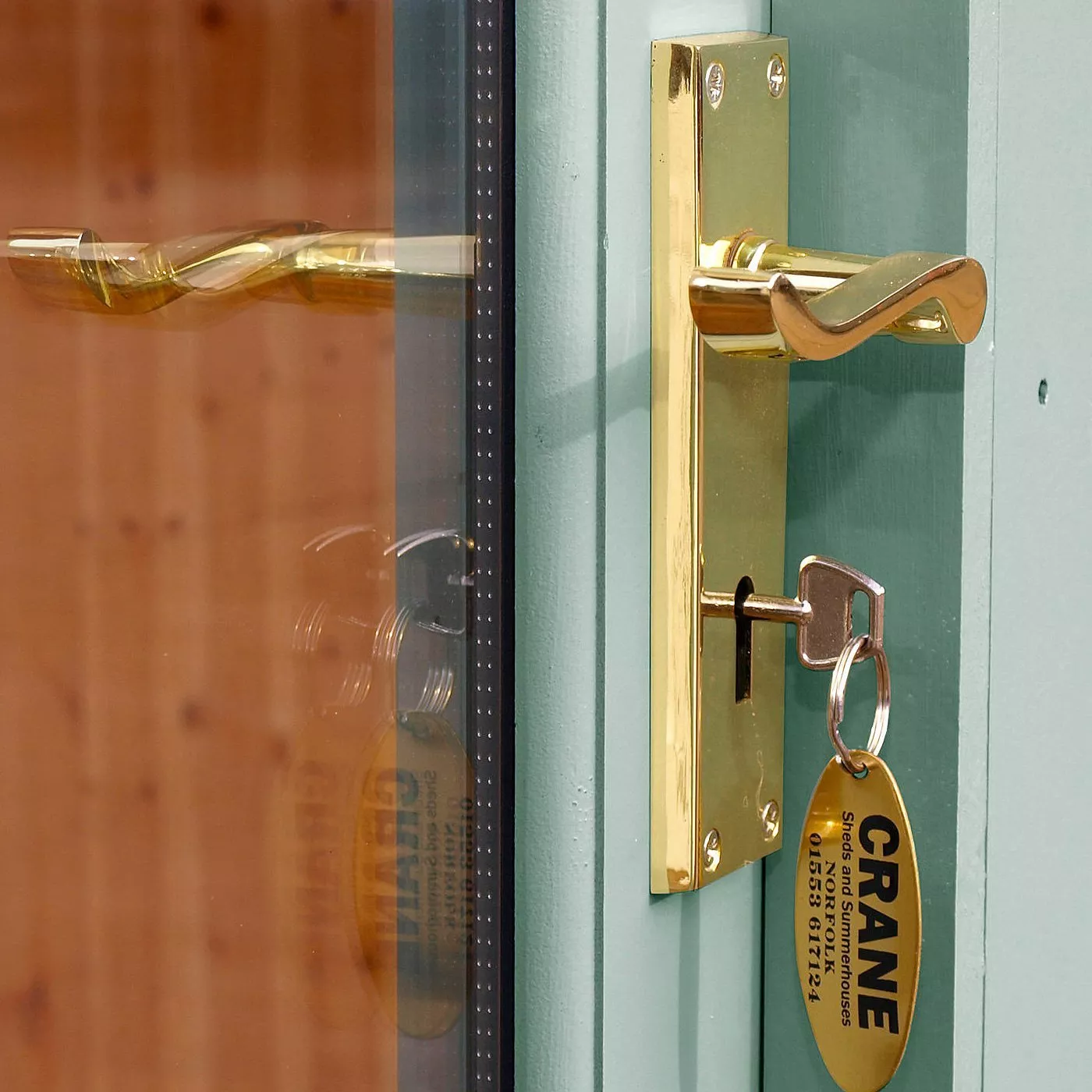 Close-up of polished brass door handle and key in lock on a teal frame, with glass panel reflection