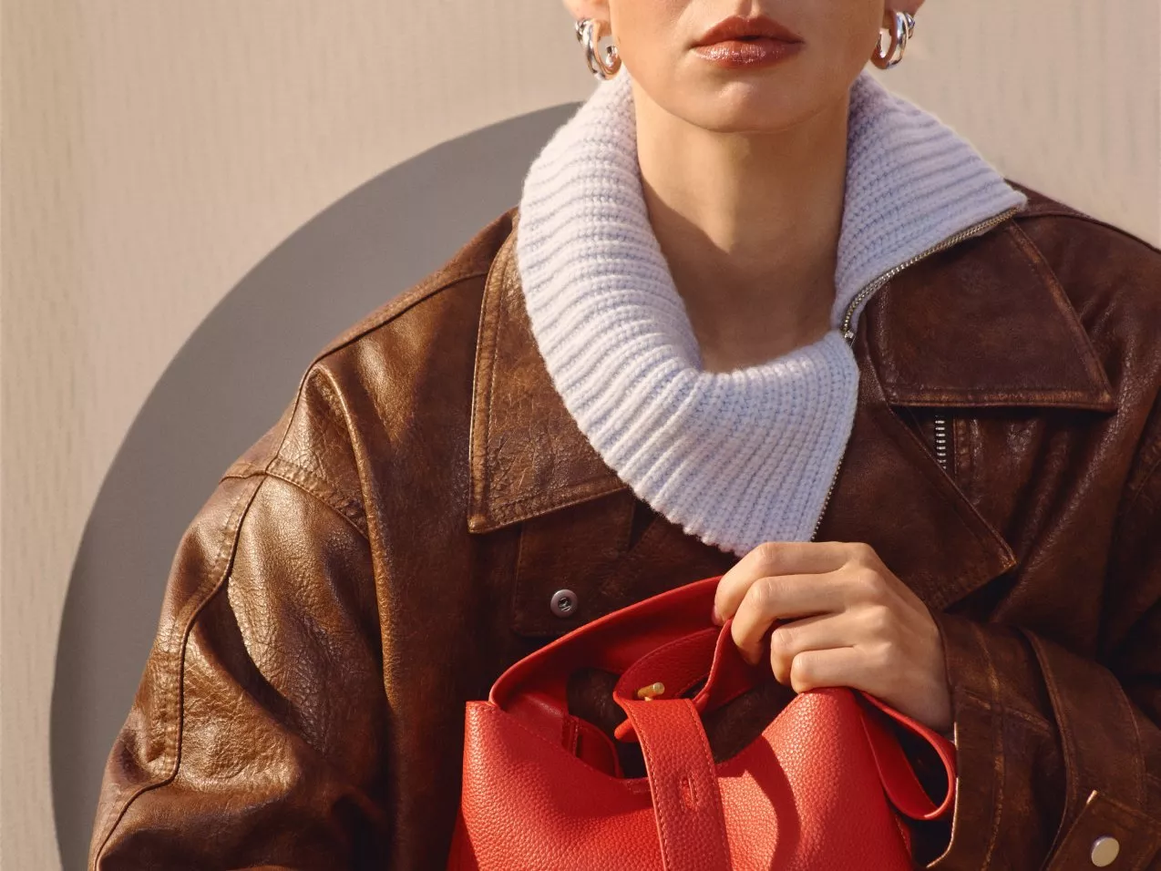 A person in a brown leather jacket and white knitwear holds a vibrant red handbag, showcasing fashion style.