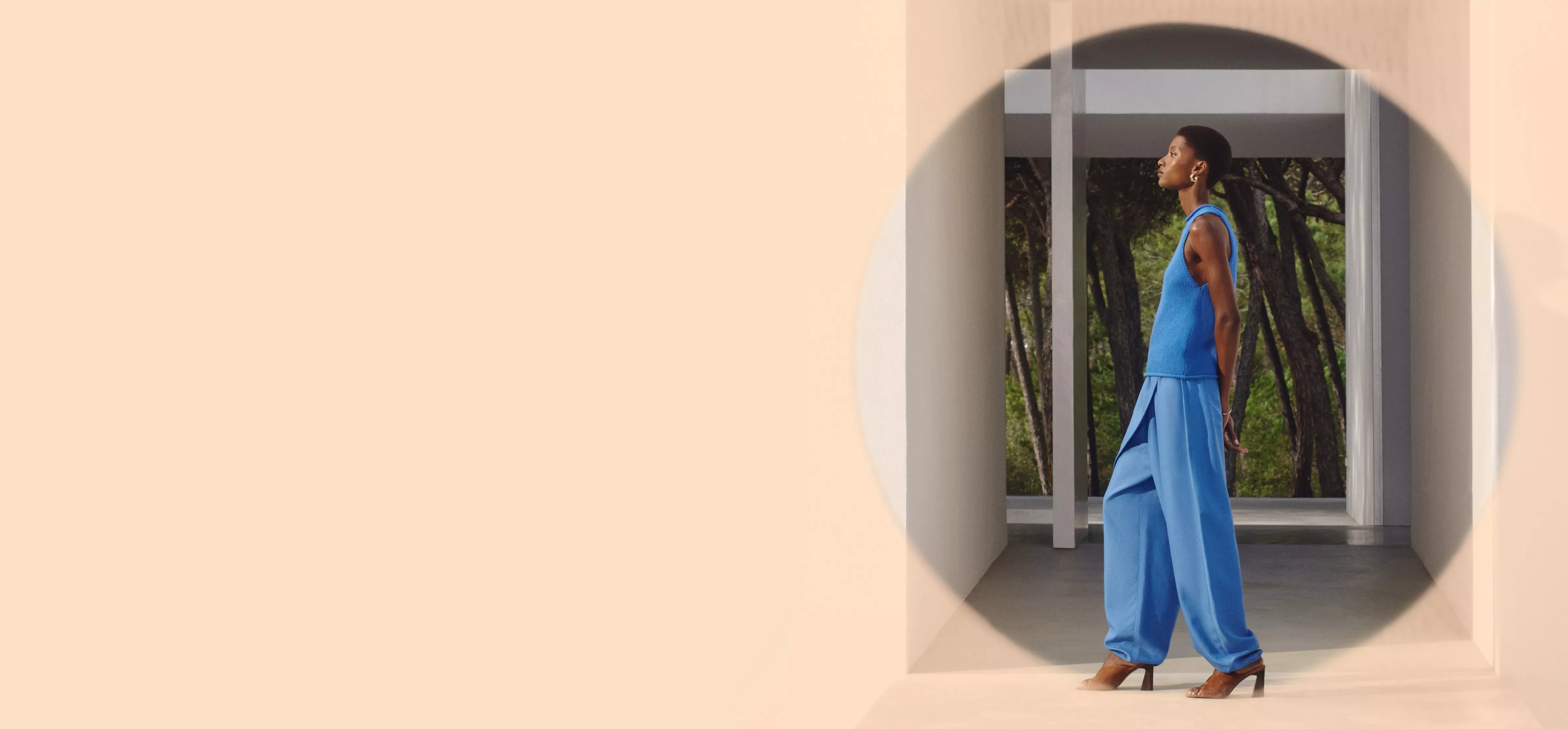 A person in a stylish blue outfit walks through a modern architectural space with trees visible in the background.