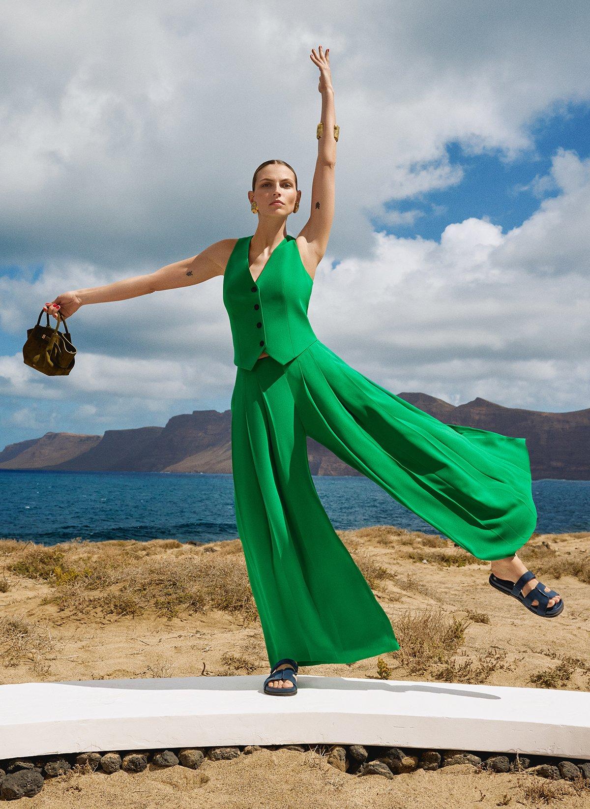 Model wearing green co-ord and black sandals, holding a handbag on the beach