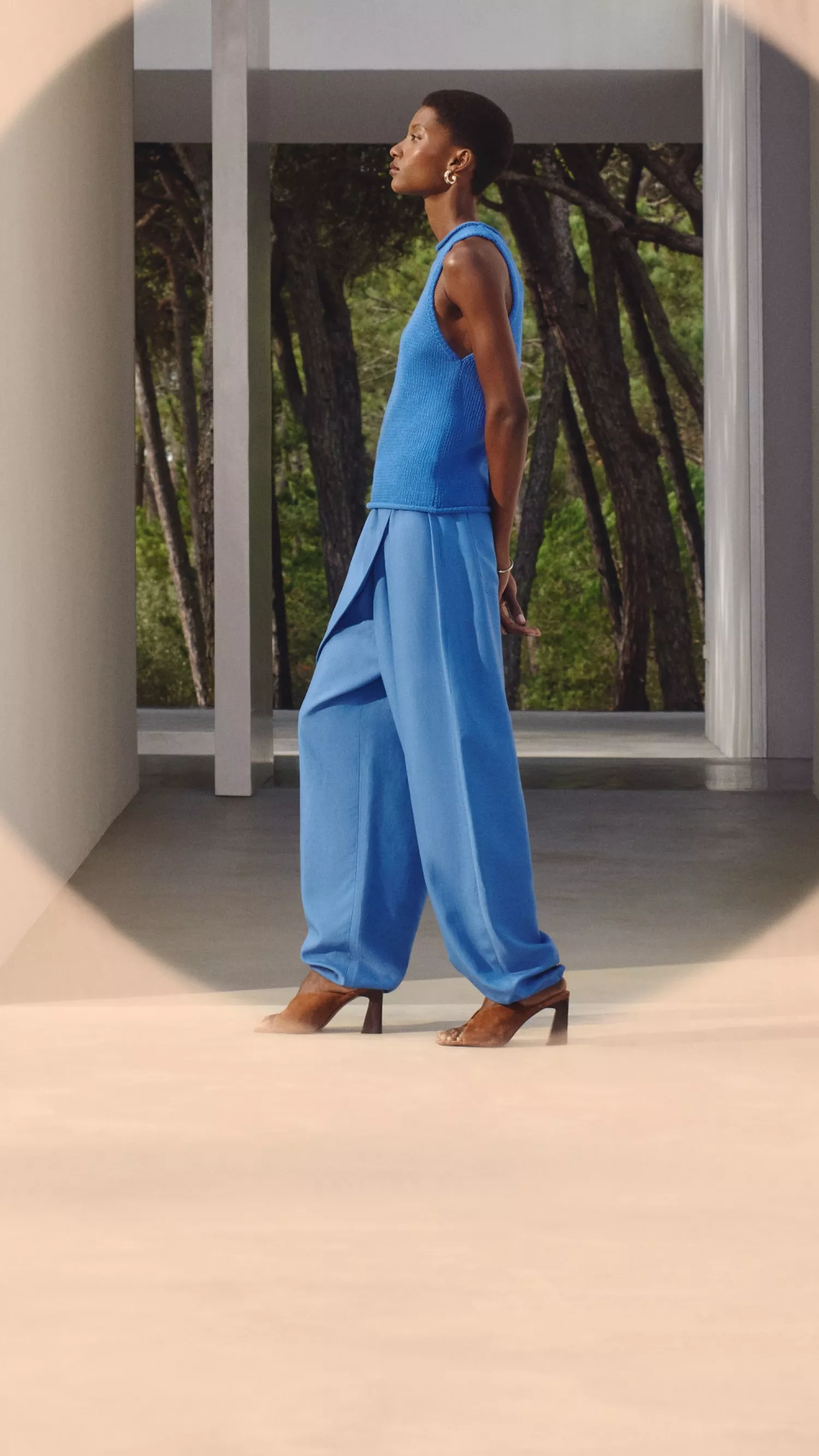 A woman in elegant blue trousers and a sleeveless top walks indoors, with a forest visible in the background.
