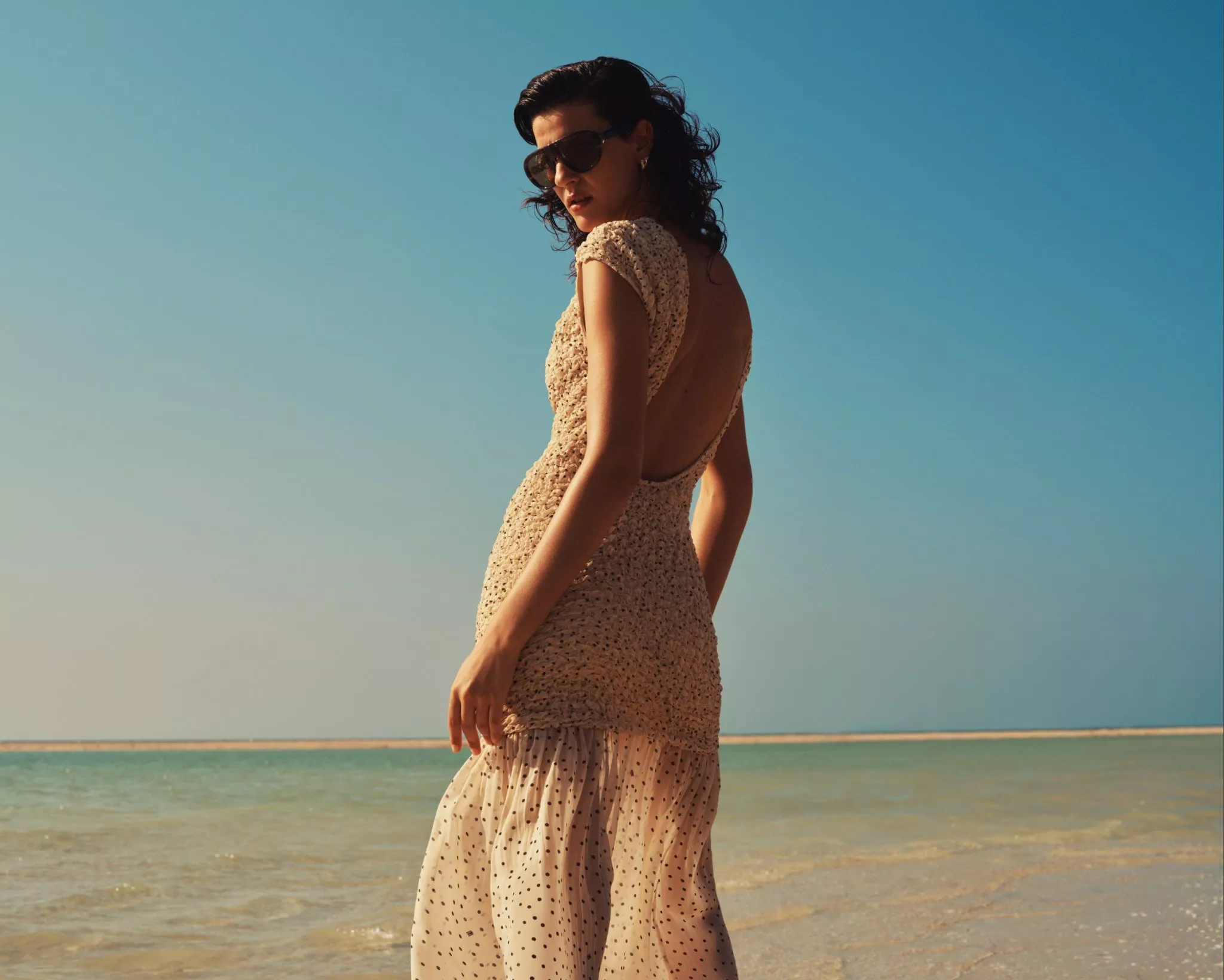 Woman in sunglasses wearing a backless summer dress on a sunny beach, with turquoise sea and clear blue sky