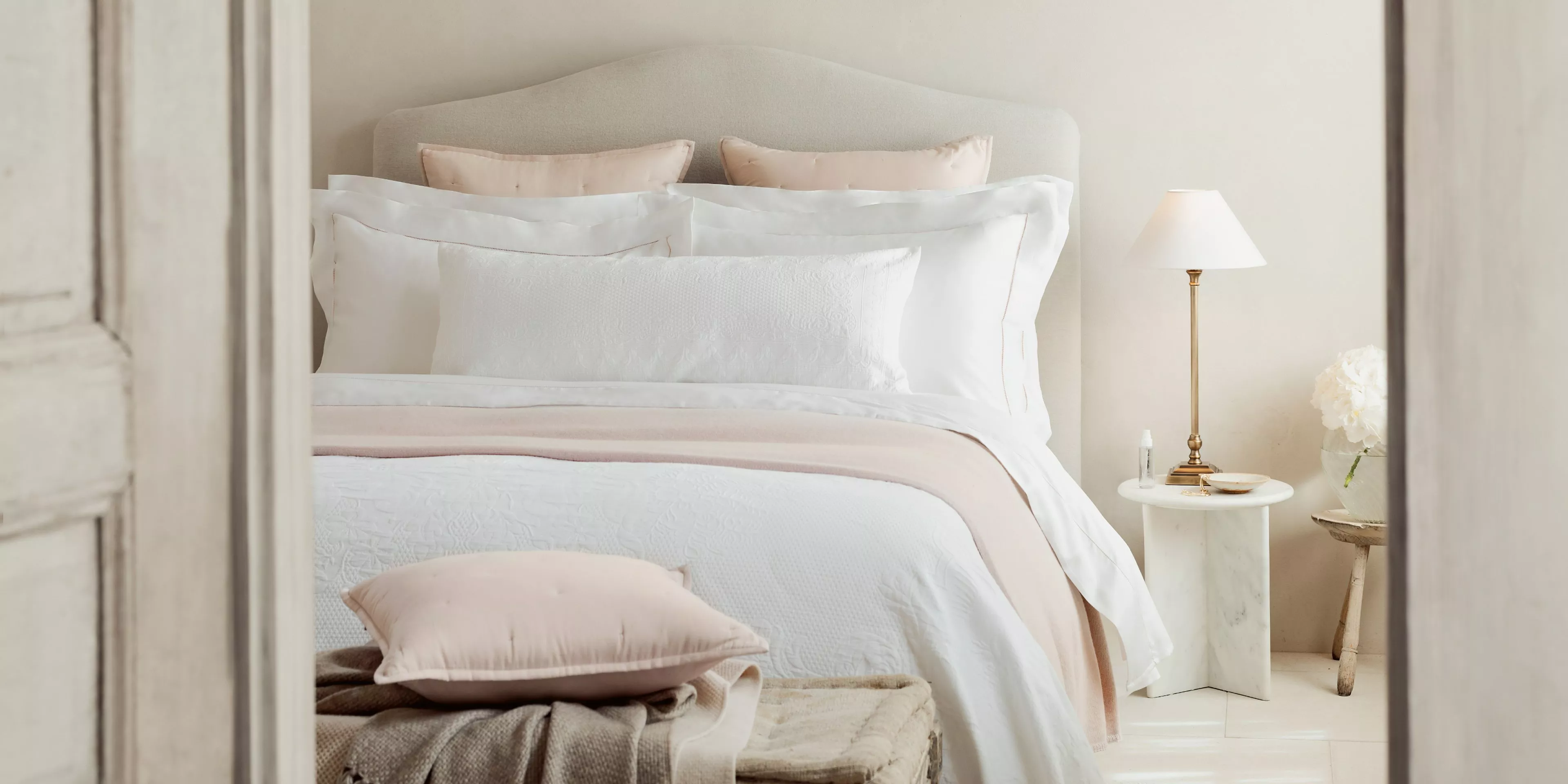 Minimalist bedroom with white bedding, upholstered headboard, and marble bedside table with lamp and candle