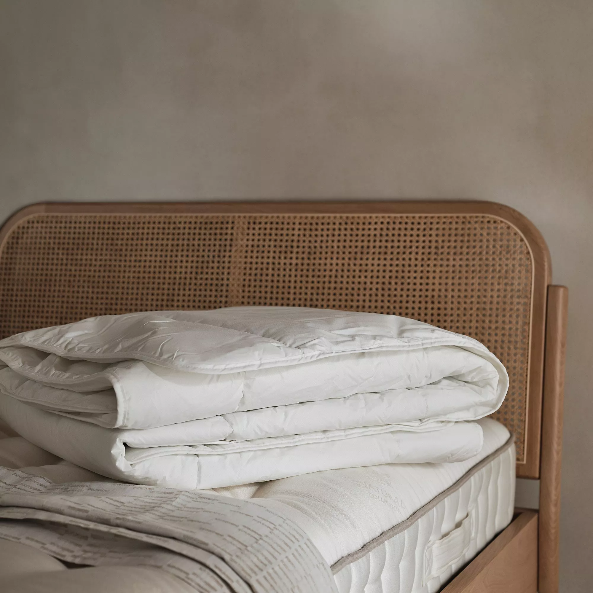 A neatly folded white duvet rests on a beige mattress with a wooden rattan headboard in a minimalist bedroom.
