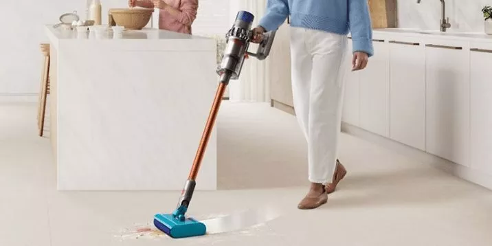 Person using cordless stick vacuum with wet roller head cleaning kitchen floor spill in modern home interior