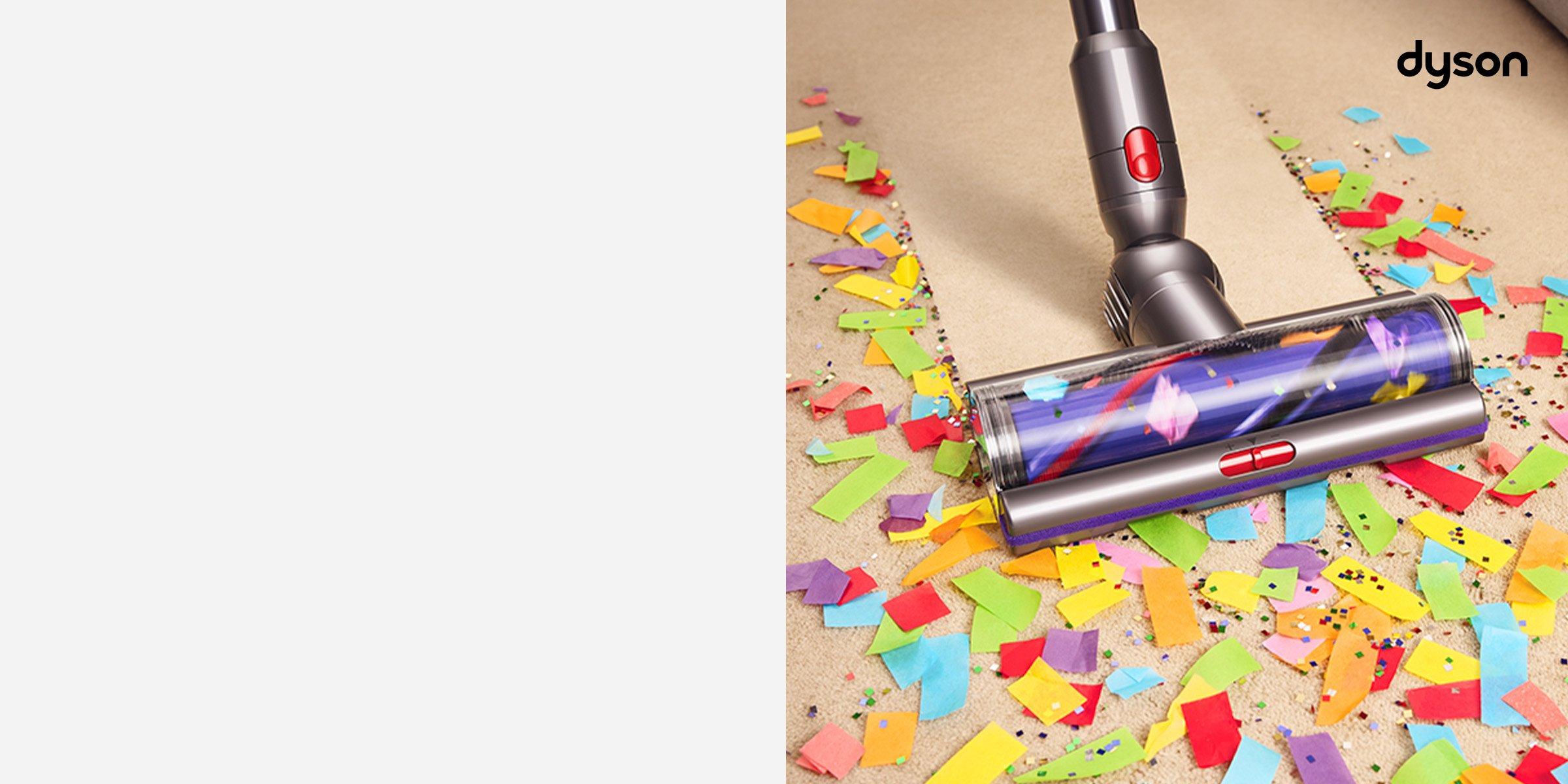 The image shows a Dyson vacuum cleaner efficiently cleaning up colorful confetti and glitter from a carpeted floor. The scene highlights the vacuum's powerful suction and cleaning capability, with a clear line showing the cleaned area versus the messy one.