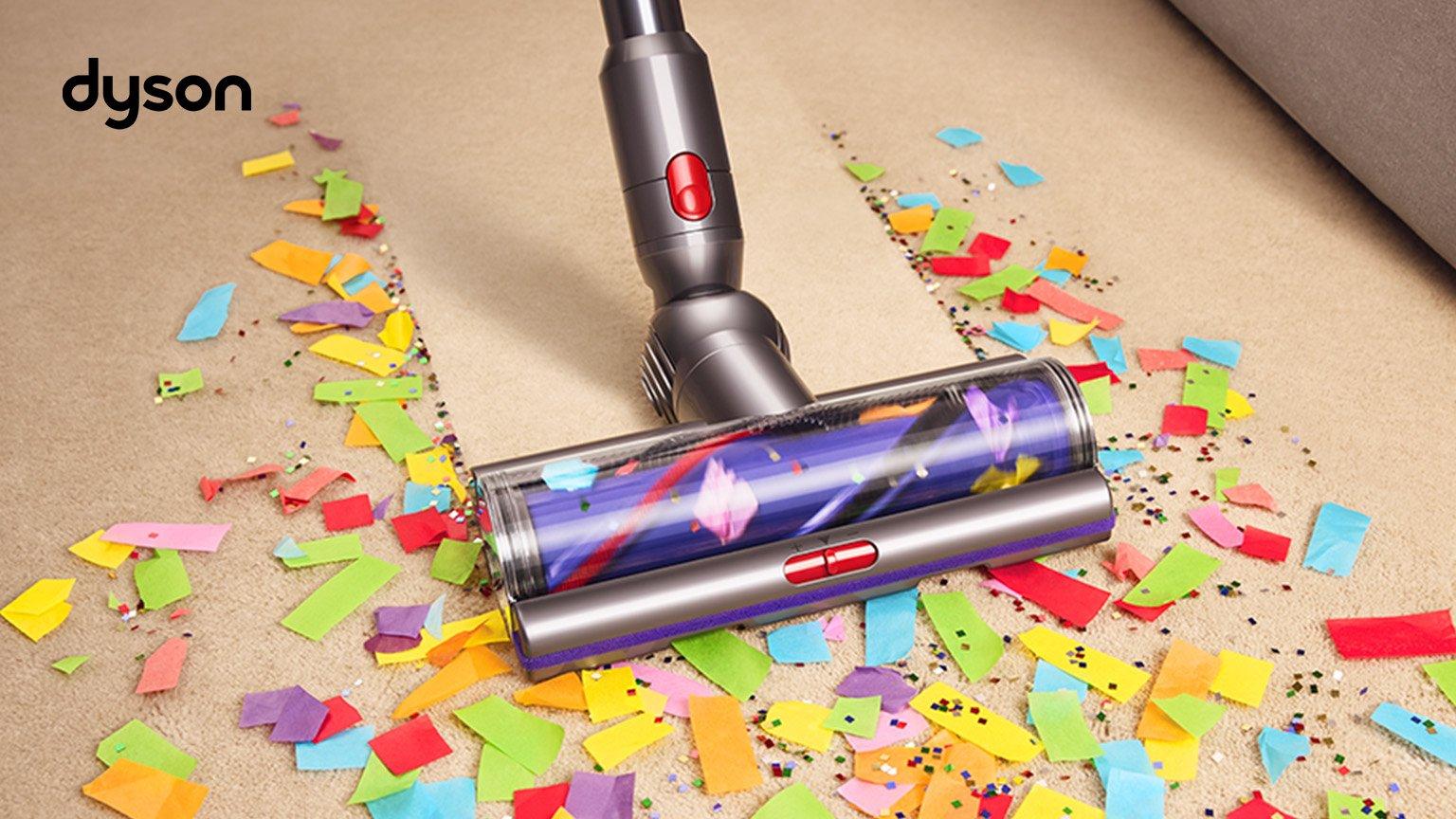 The image shows a Dyson vacuum cleaner efficiently cleaning up colorful confetti and glitter from a carpeted floor. The scene highlights the vacuum's powerful suction and cleaning capability, with a clear line showing the cleaned area versus the messy one.