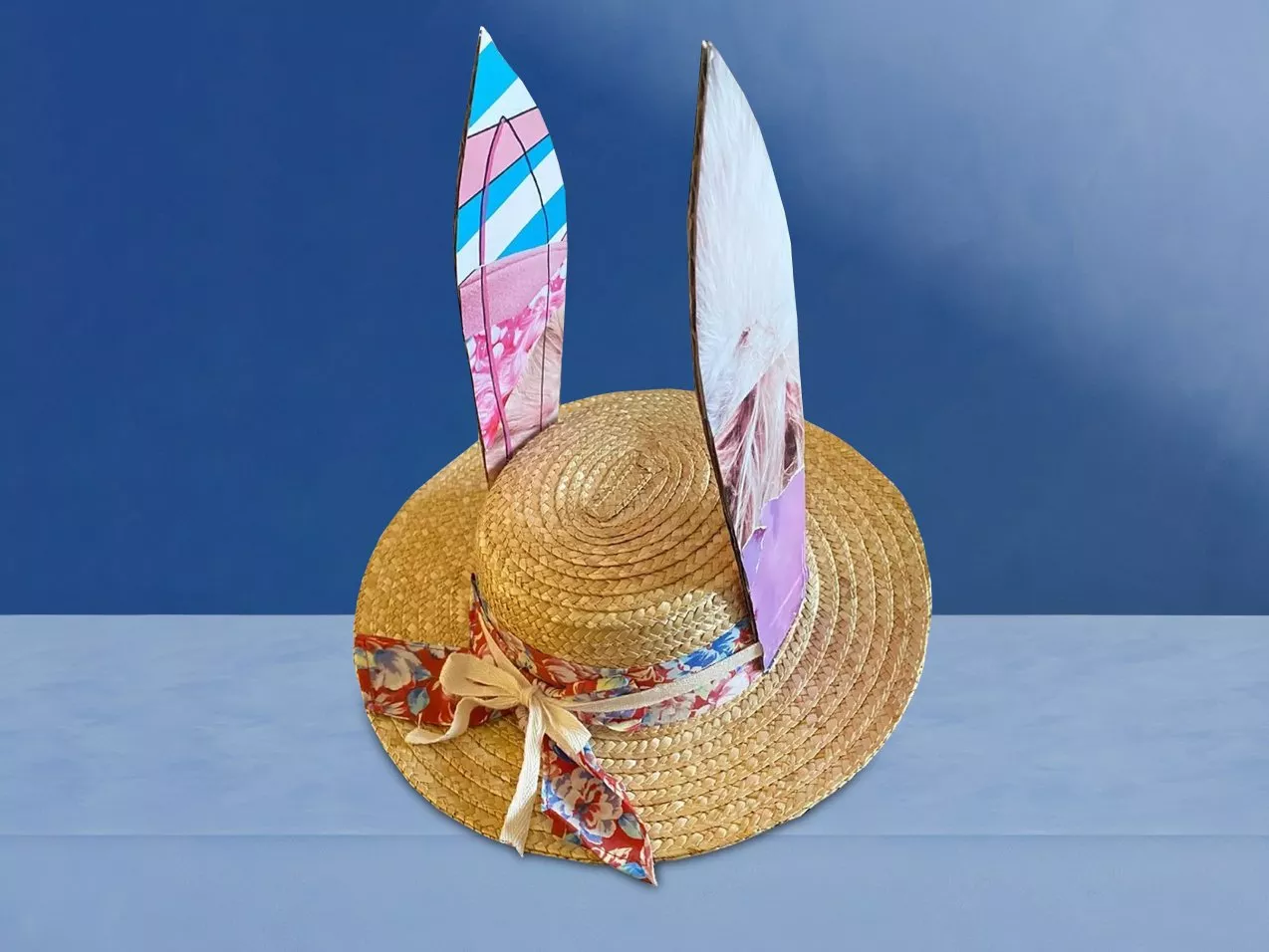 A straw hat with colourful, patterned ears and a floral ribbon sits on a blue surface against a blue background.