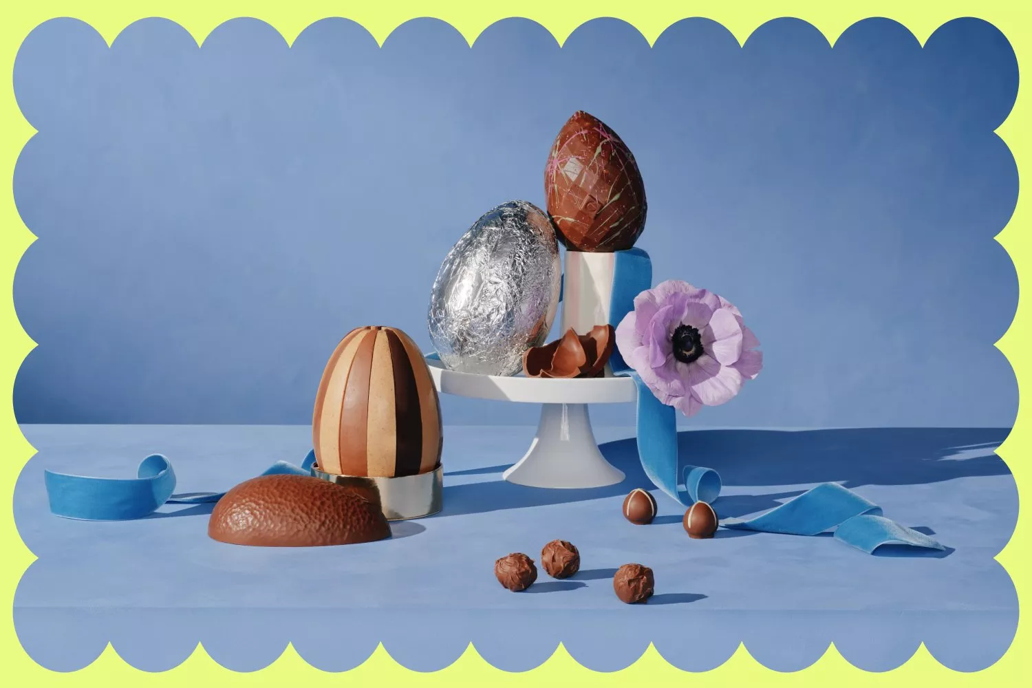 Elegant display of chocolate eggs on a stand, alongside a purple flower, blue ribbon, and scattered chocolates.