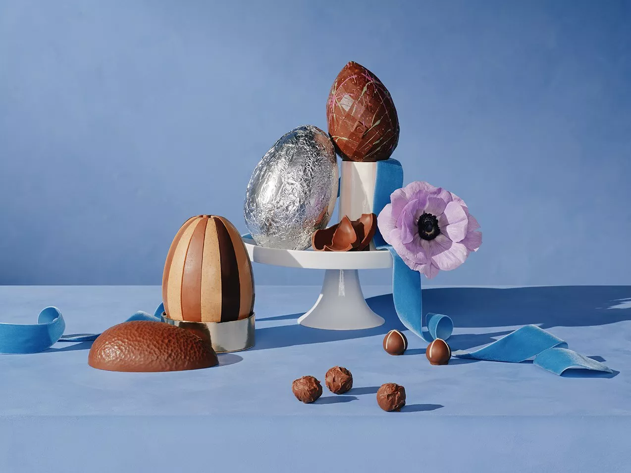 A variety of chocolate eggs and truffles displayed on a blue table. Includes a purple flower and blue ribbon.