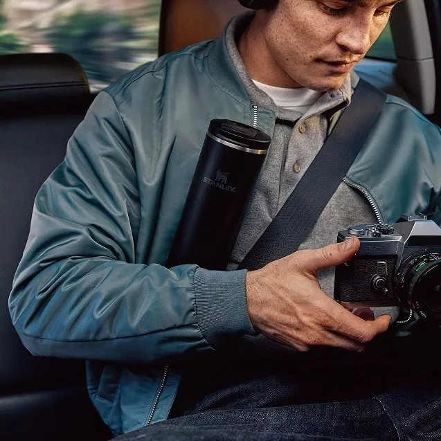Man in a car wearing headphones, holding a vintage camera and a Stanley travel mug. Casual clothing suggests travel.