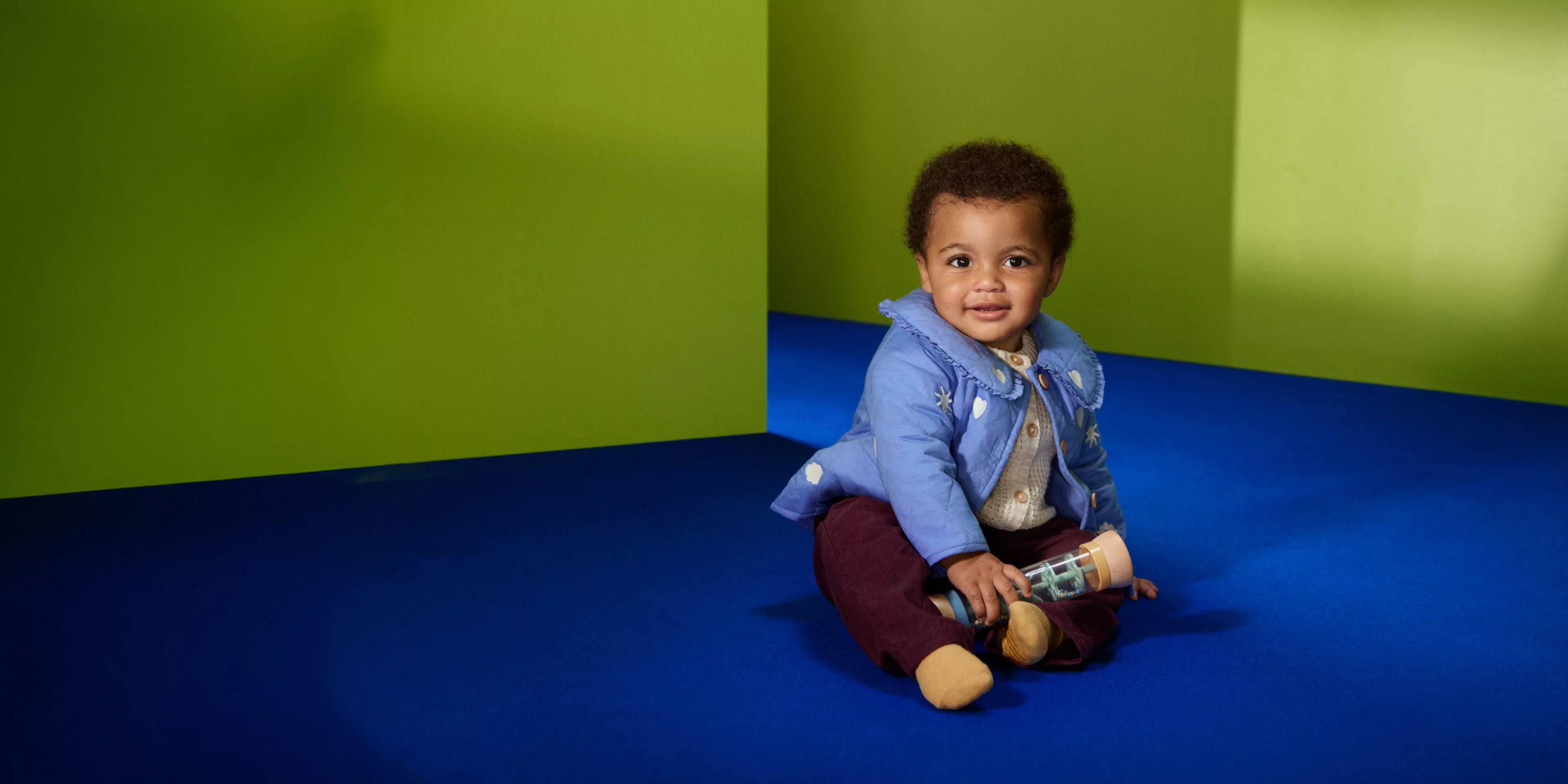 A toddler in a yellow outfit with white duck patterns sits on a blue rug next to greenery, looking upwards.