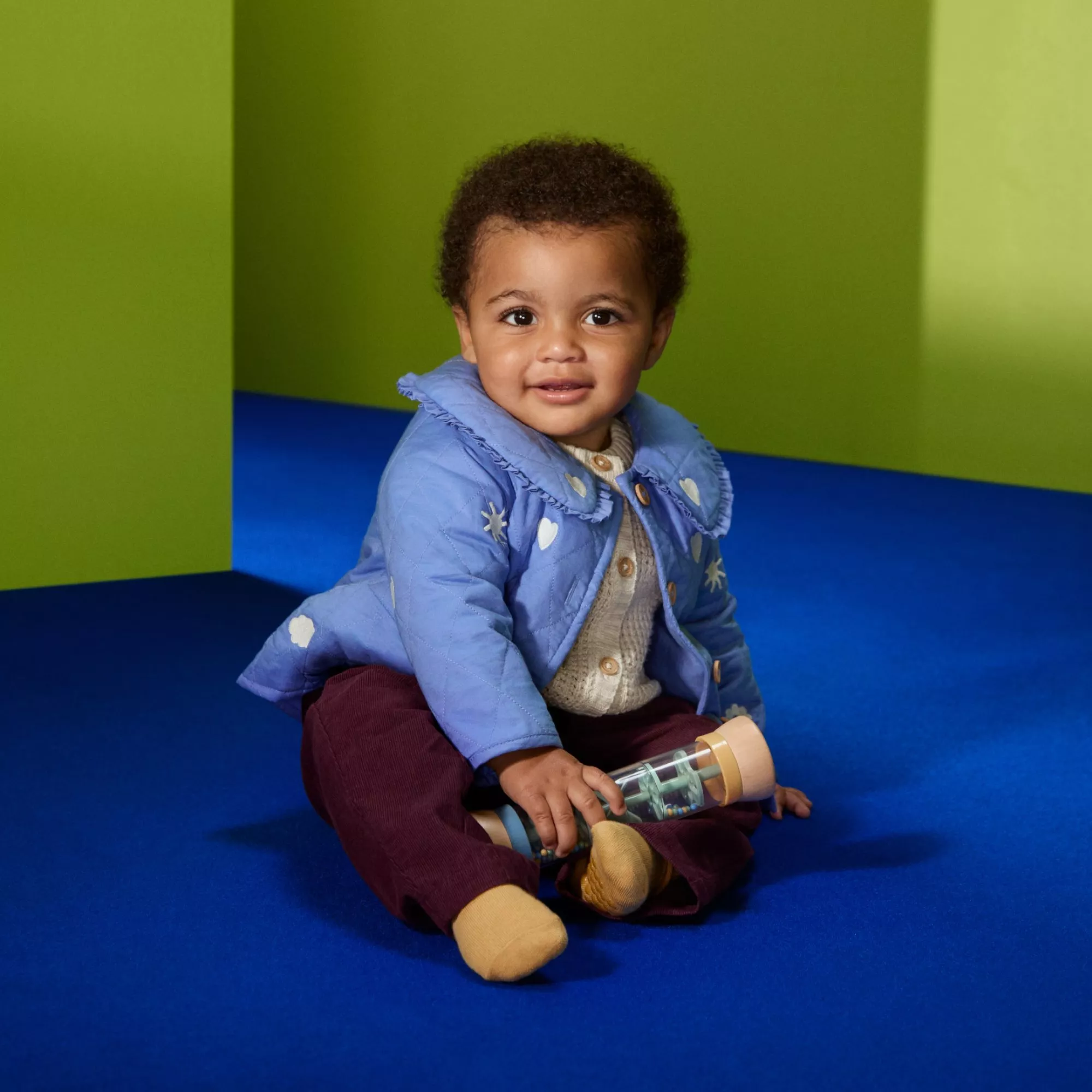 Smiling toddler in blue jacket sitting on blue floor, holding toy bottle in bright green playroom setting.
