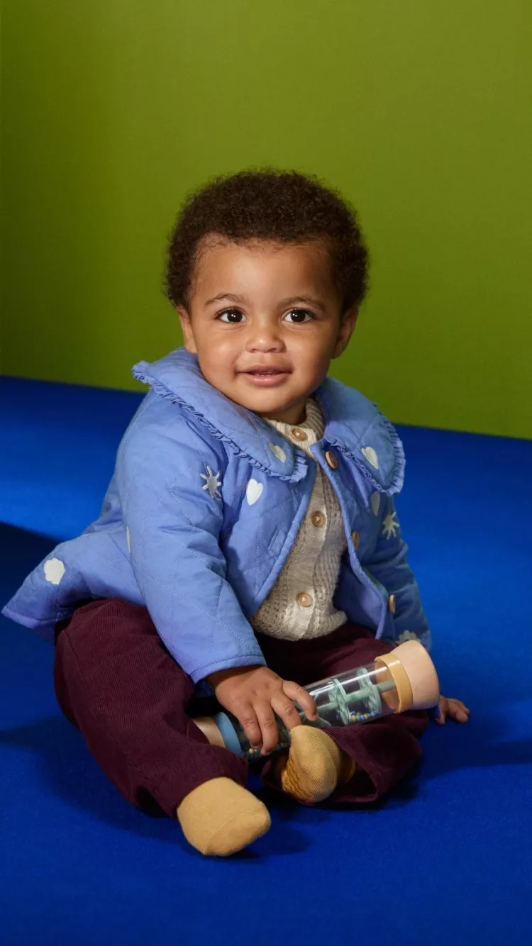 A baby wearing a yellow knit outfit with duck patterns sits on a blue carpet, surrounded by green foliage.