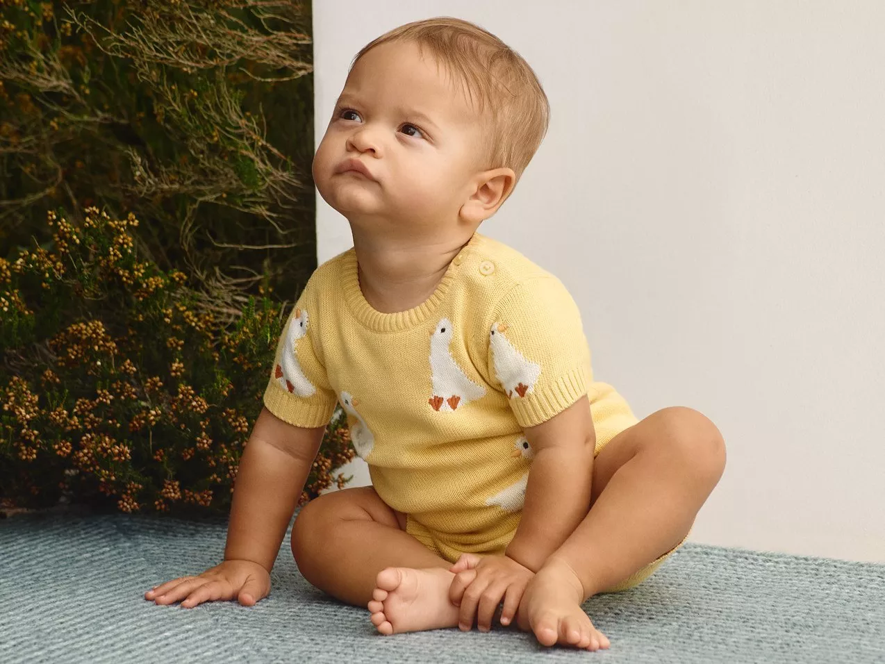 A baby in a yellow outfit with duck prints sits on a blue carpet, looking upwards thoughtfully near greenery.